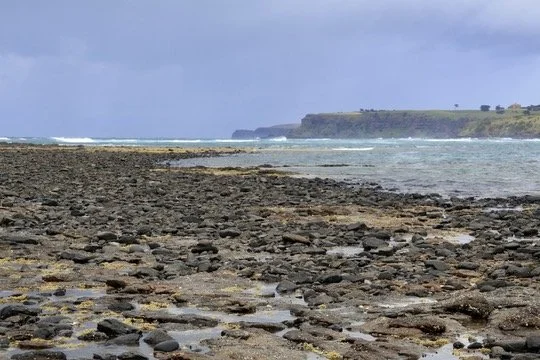 Rocky shoreline with ocean waves and a distant green cliff under a cloudy sky.