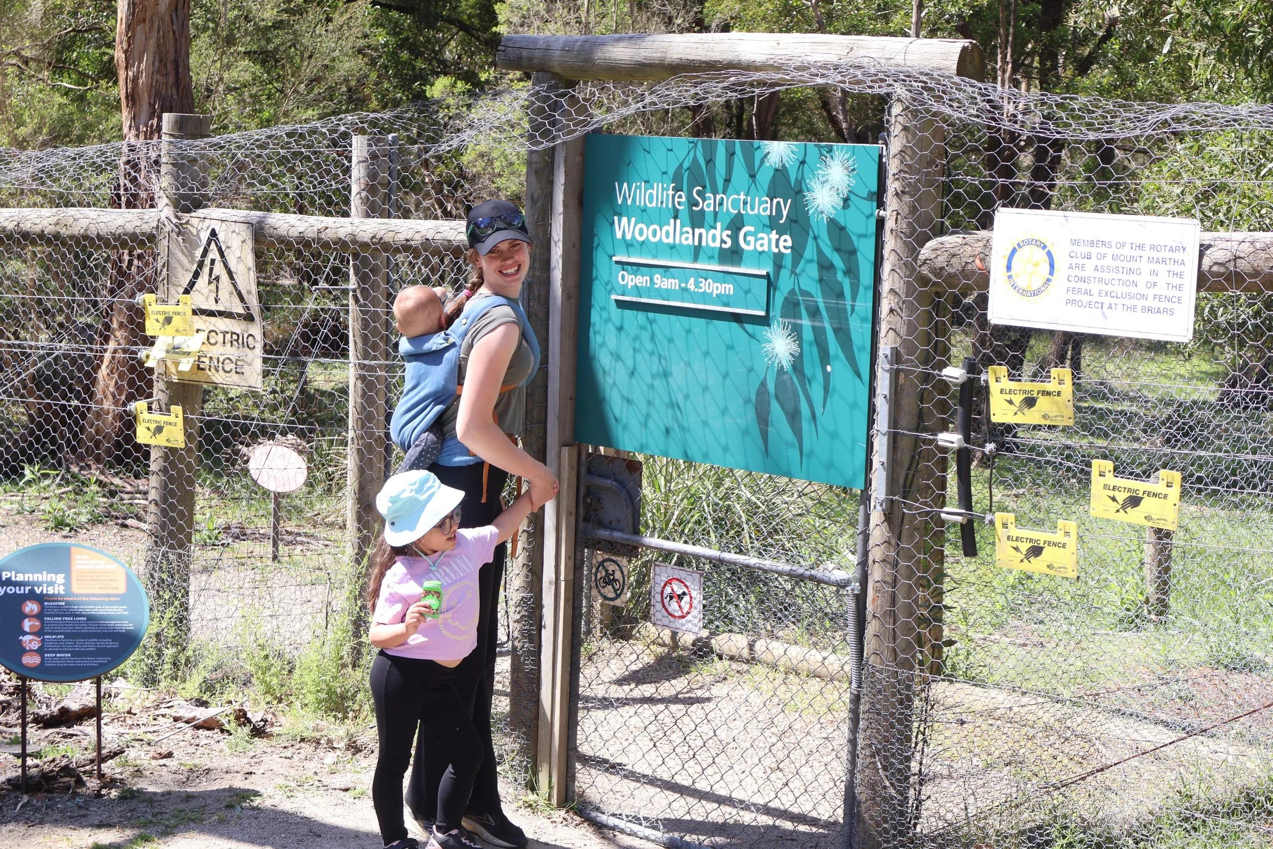 A woman and two children standing at the entrance of a wildlife sanctuary, with the woman carrying a baby in a backpack carrier. They are smiling and holding a sign near the wildlife sanctuary gate, which has a large teal sign that reads 'Wildlife Sanctuary Woodlands Gate' with open hours listed. The gate is surrounded by a wire fence with yellow signs that say 'Electric Fence'. There are trees and greenery in the background.