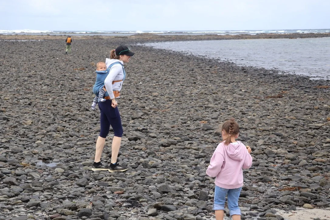 A woman carrying a baby on her back walking on a rocky beach with a girl in a pink hoodie and blue jeans nearby, and another person in the distance by the water.