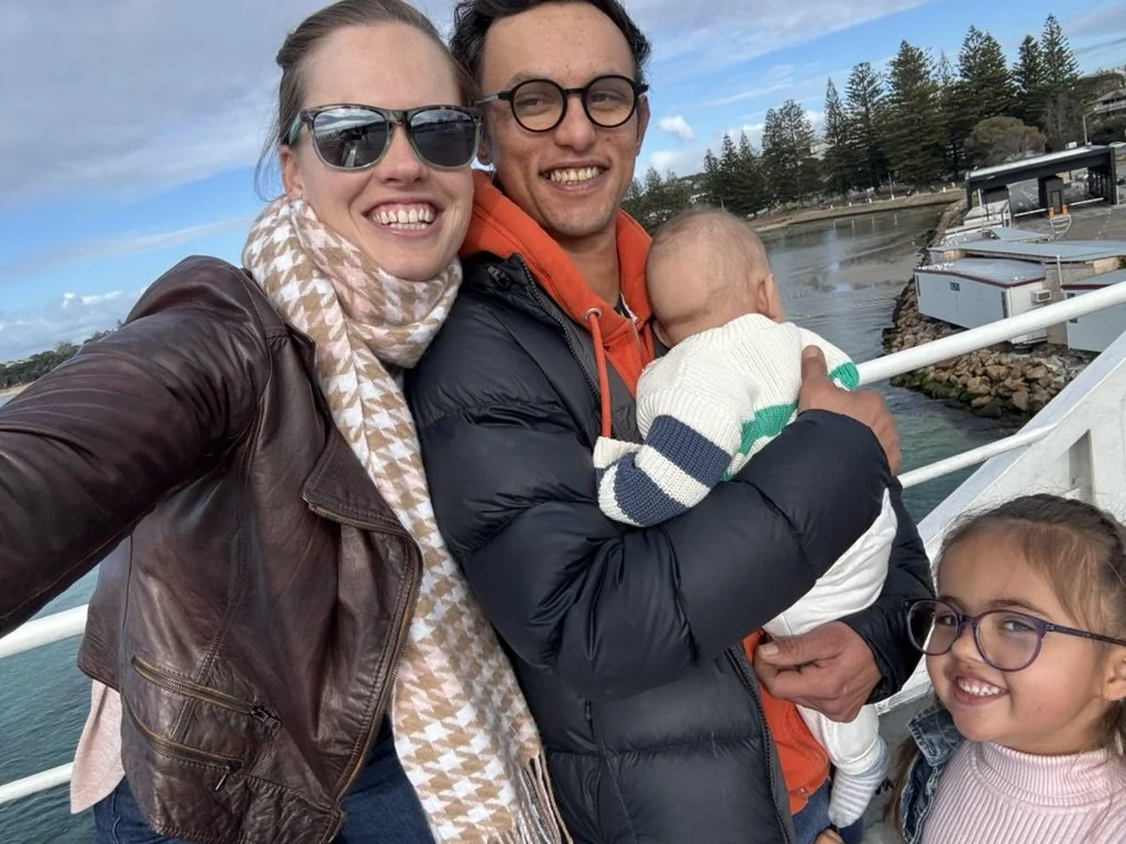 A smiling family of four, including a woman, a man holding a baby, and a young girl, taking a selfie outdoors near water with boats and trees in the background.