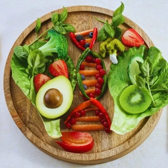 A wooden plate decorated with various sliced fruits and vegetables arranged to resemble a human face, including an avocado for the nose, strawberries, kiwi, cherry tomatoes, cucumbers, broccoli, carrots, and bell peppers.