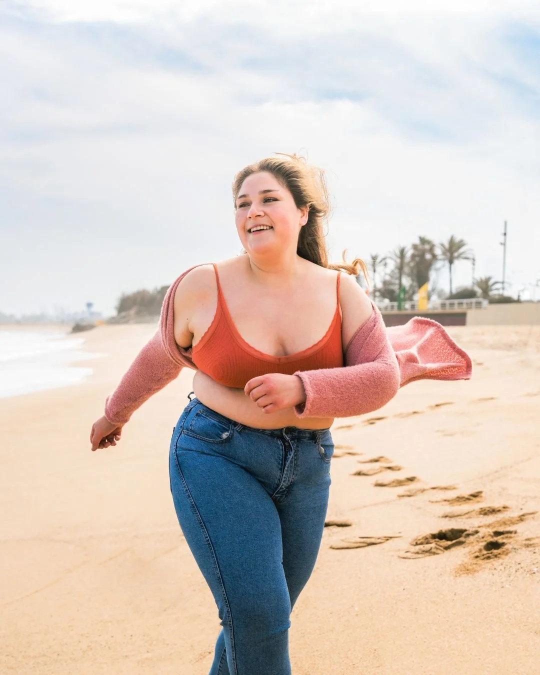 A woman running on the beach, smiling, wearing a red top and a pink cardigan with jeans, with a cloudy sky and palm trees in the background.