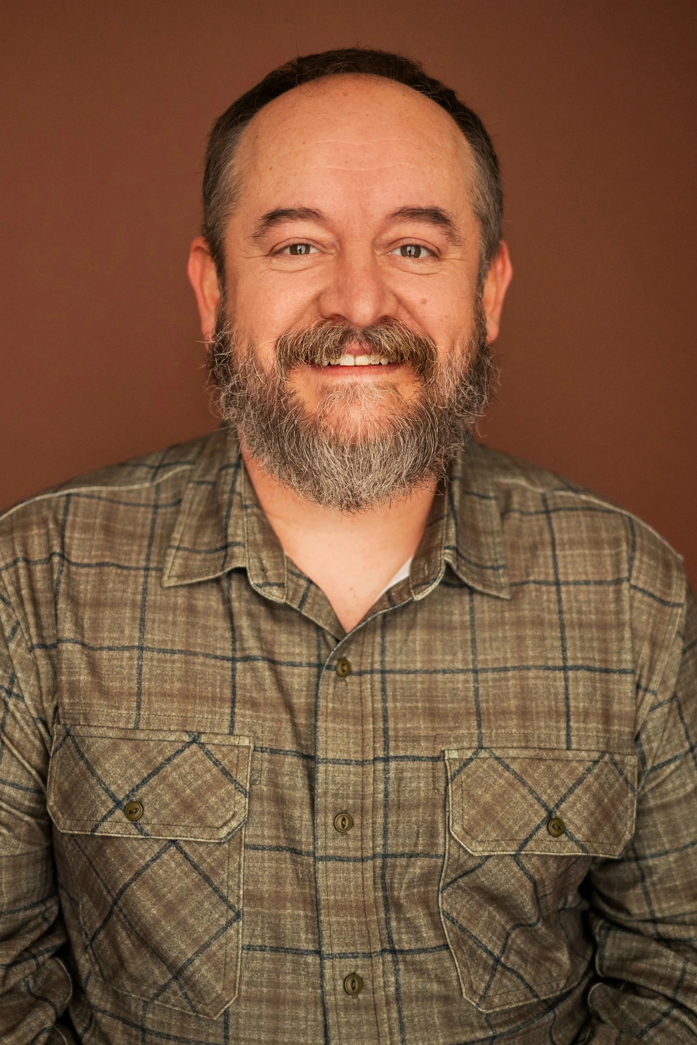 A man with a beard and mustache smiling, wearing a plaid shirt, in front of a brown background.