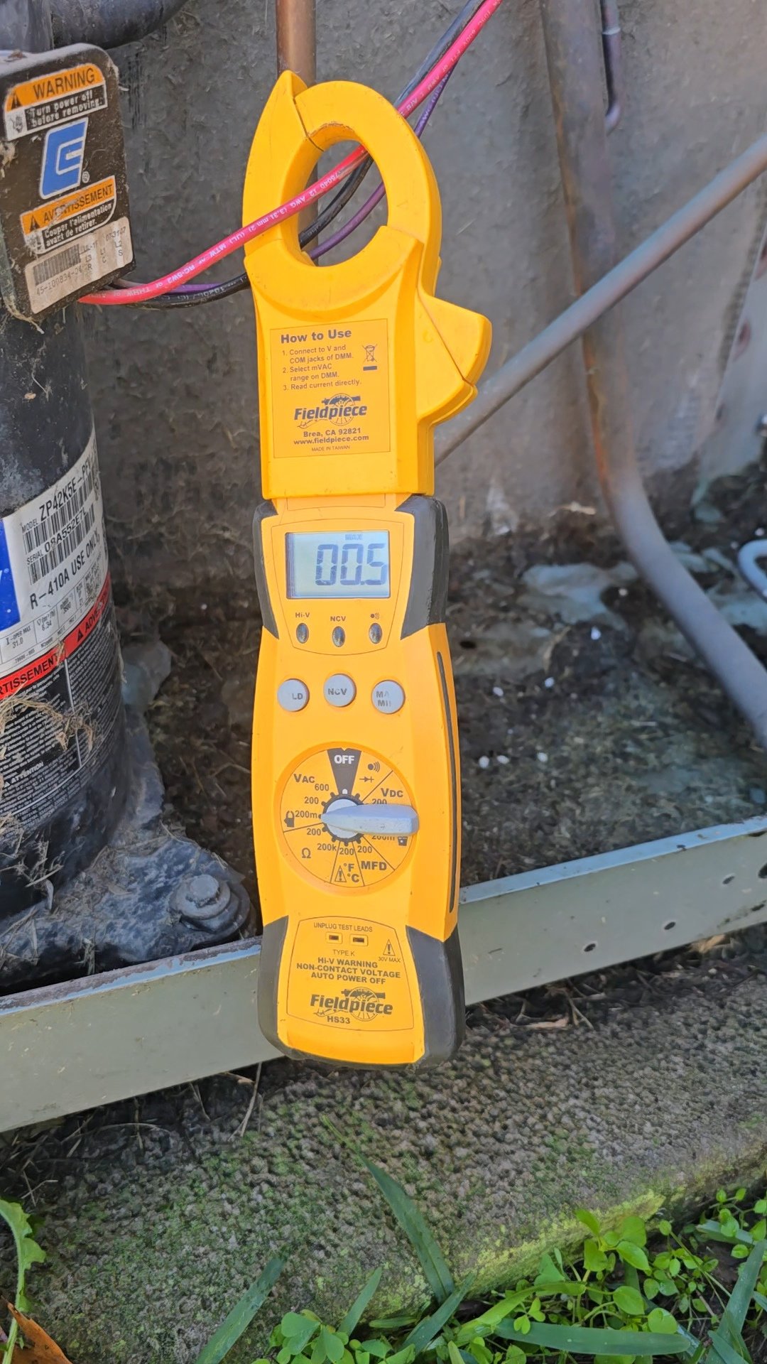 A yellow digital multimeter is measuring voltage near an electrical device, displaying 0.05 on its screen, outdoors next to some wires and equipment.