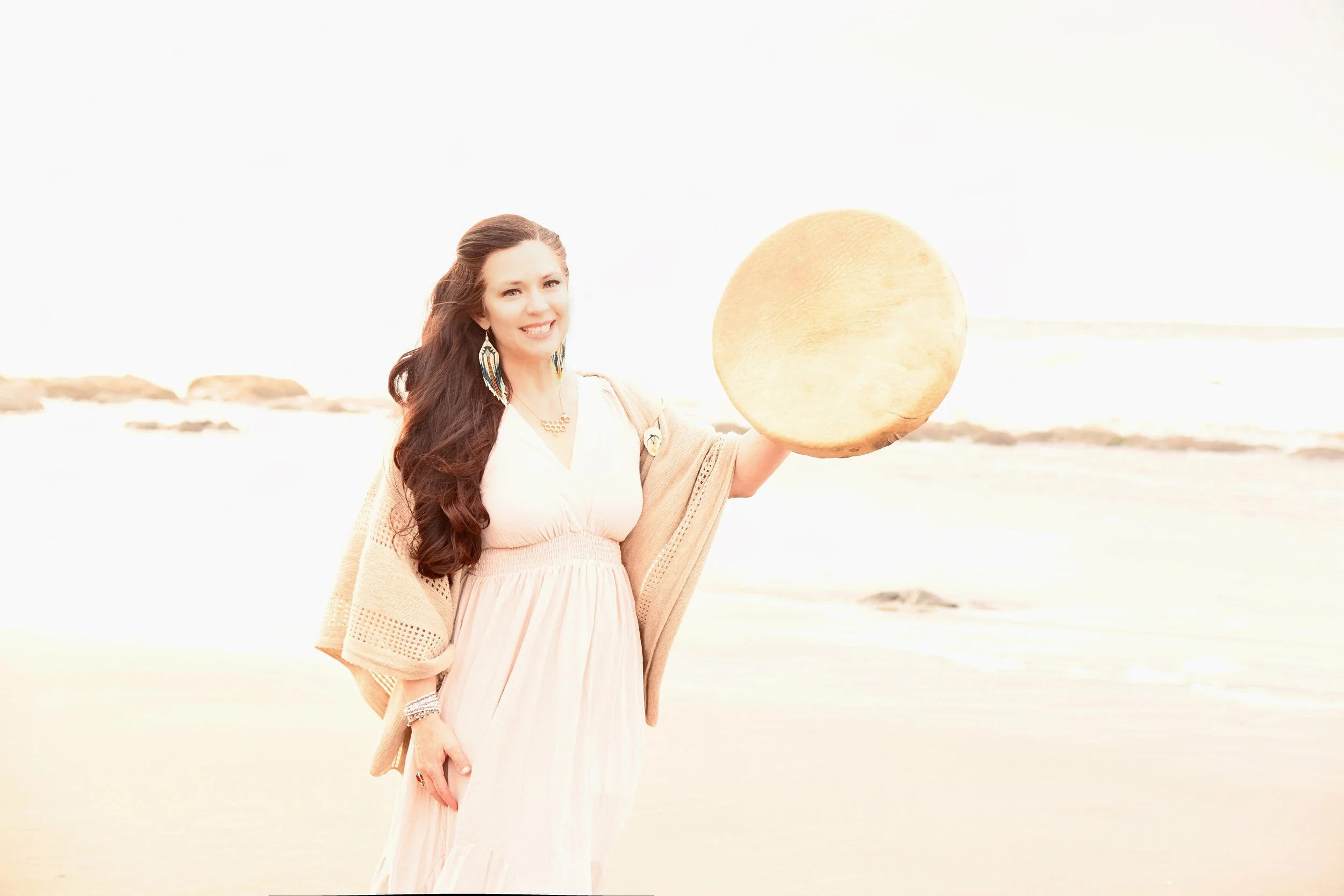 A woman holding a large tambourine on a beach with bright sunlight.