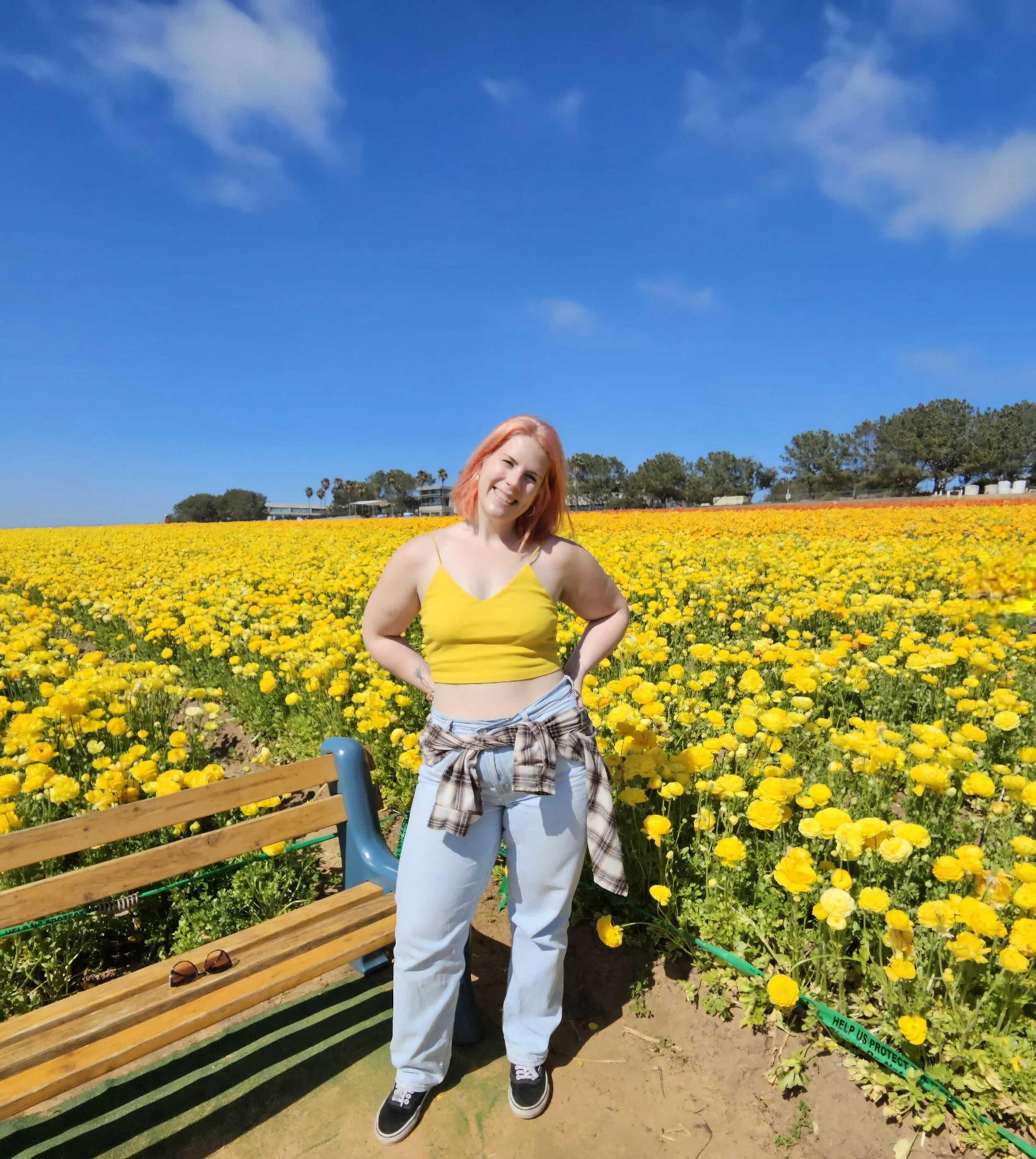 A woman with pink hair in a yellow crop top and white pants standing in a field of yellow flowers under a blue sky