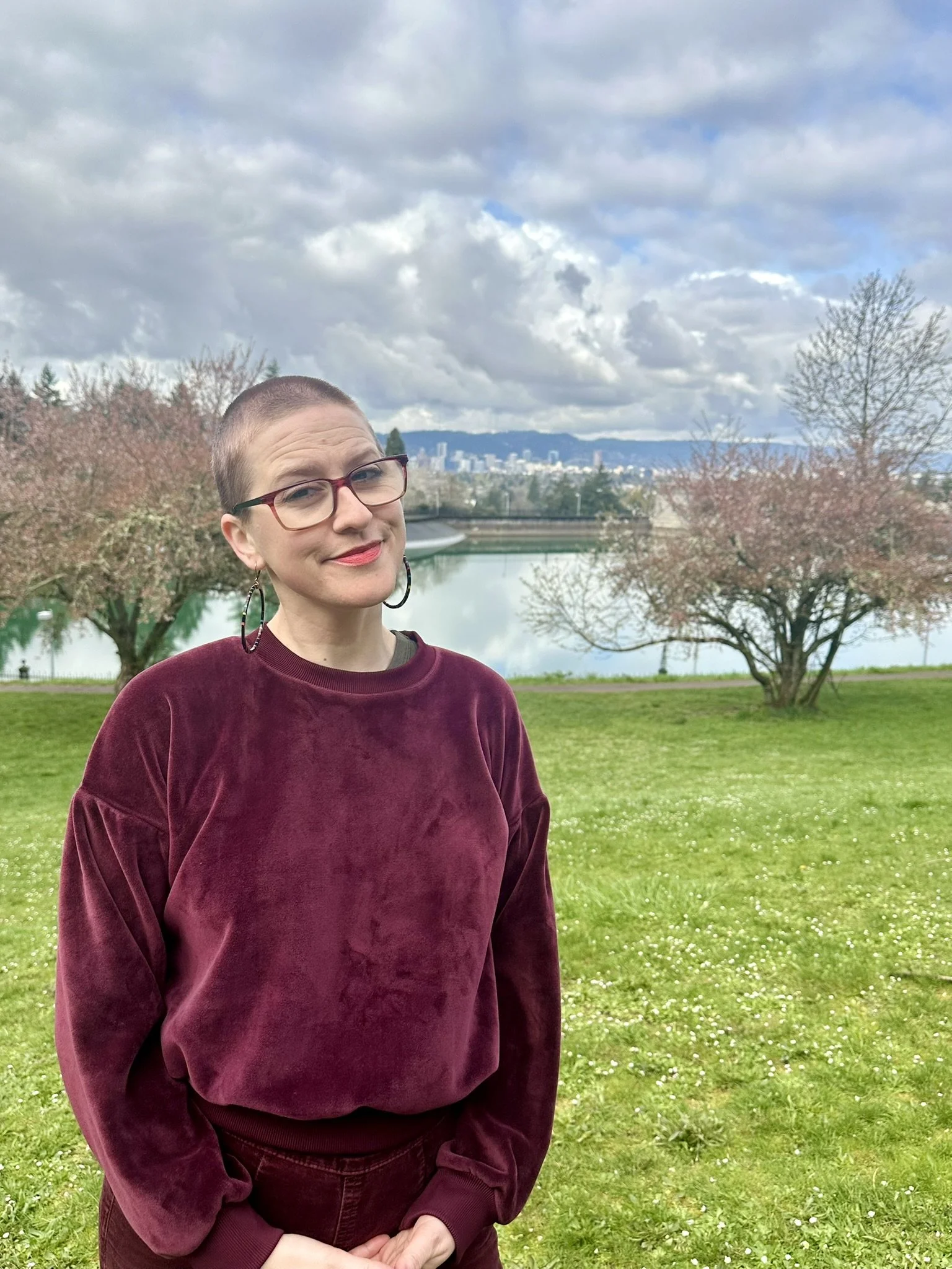 A woman with short hair, glasses, and hoop earrings standing outdoors in a park near the water, with trees and city skyline in the background.