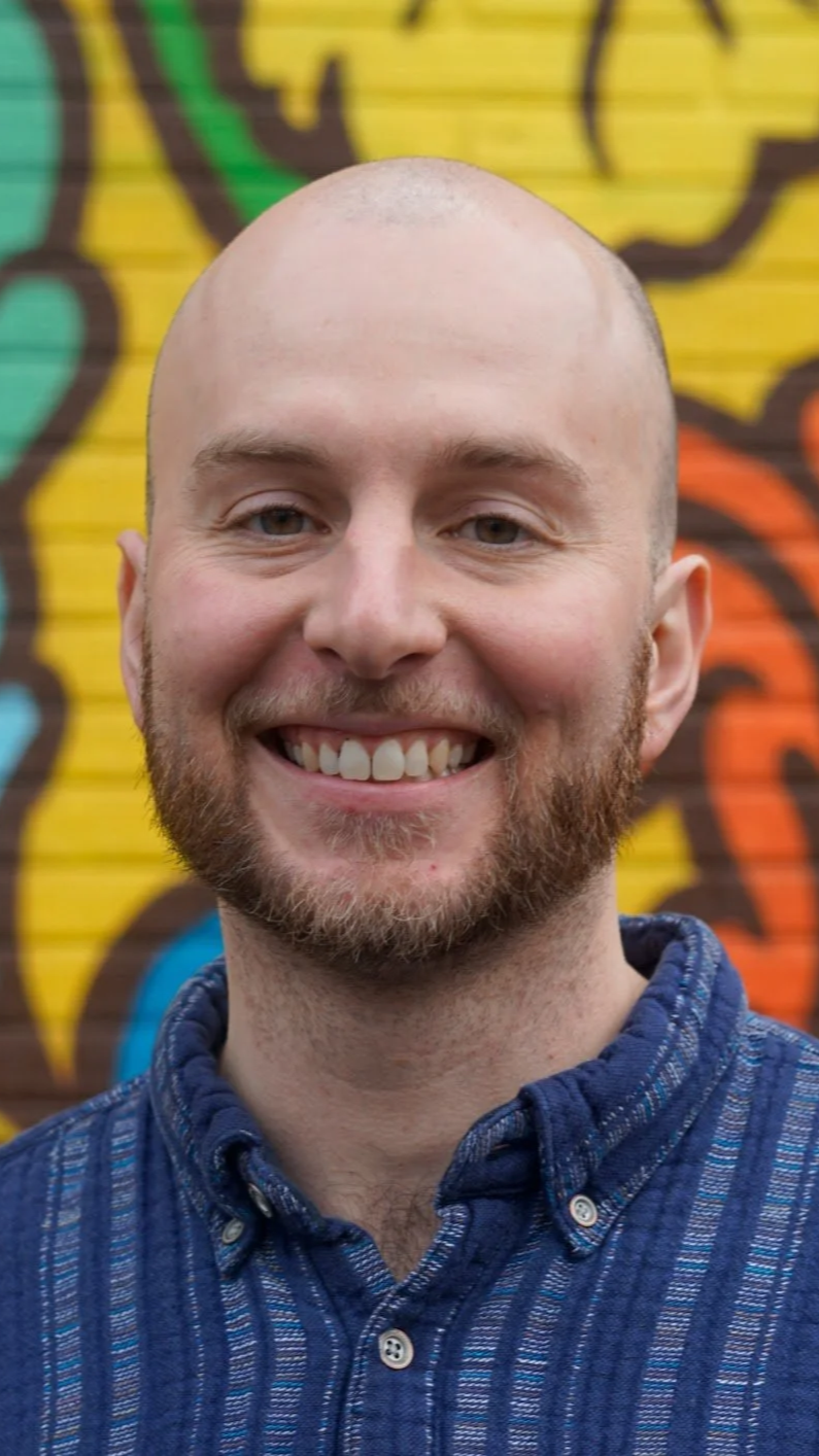 A smiling man with a bald head and a beard standing in front of a colorful graffiti wall, wearing a blue plaid button-up shirt.