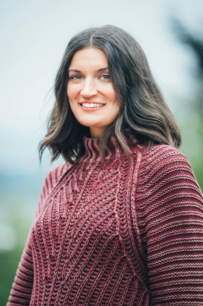 A woman with shoulder-length wavy dark hair, smiling, wearing a maroon knitted sweater outdoors with blurred greenery in the background.