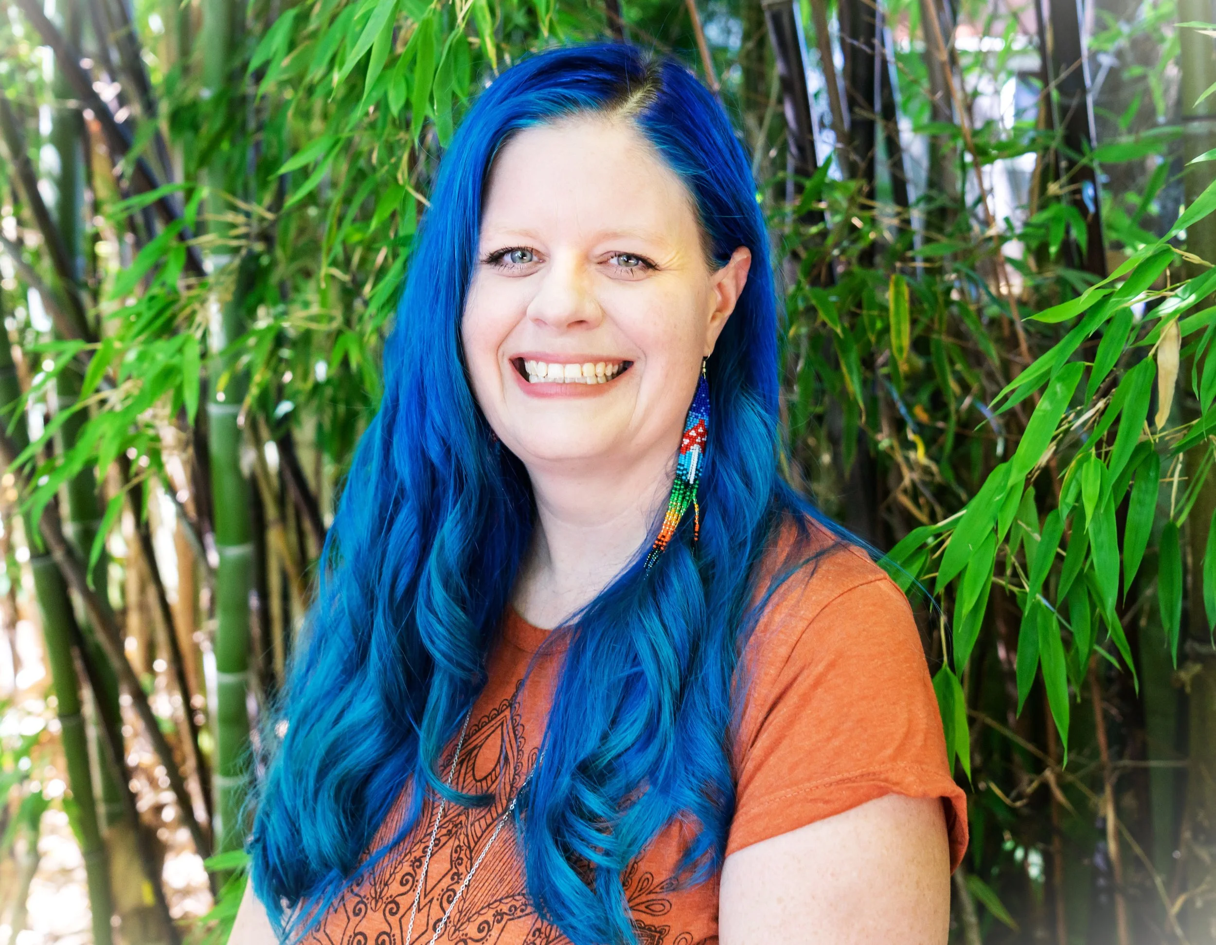 A woman with long bright blue hair smiling outdoors with green bamboo plants in the background.