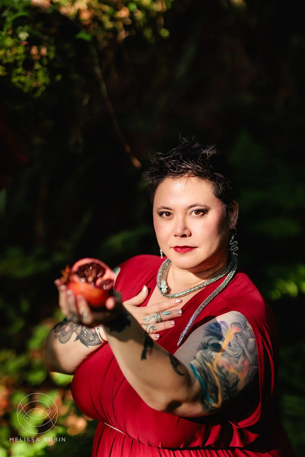 A woman with short dark hair and tattoos on her arms, wearing a red dress and jewelry, is holding a pomegranate in an outdoor setting with blurred greenery in the background.