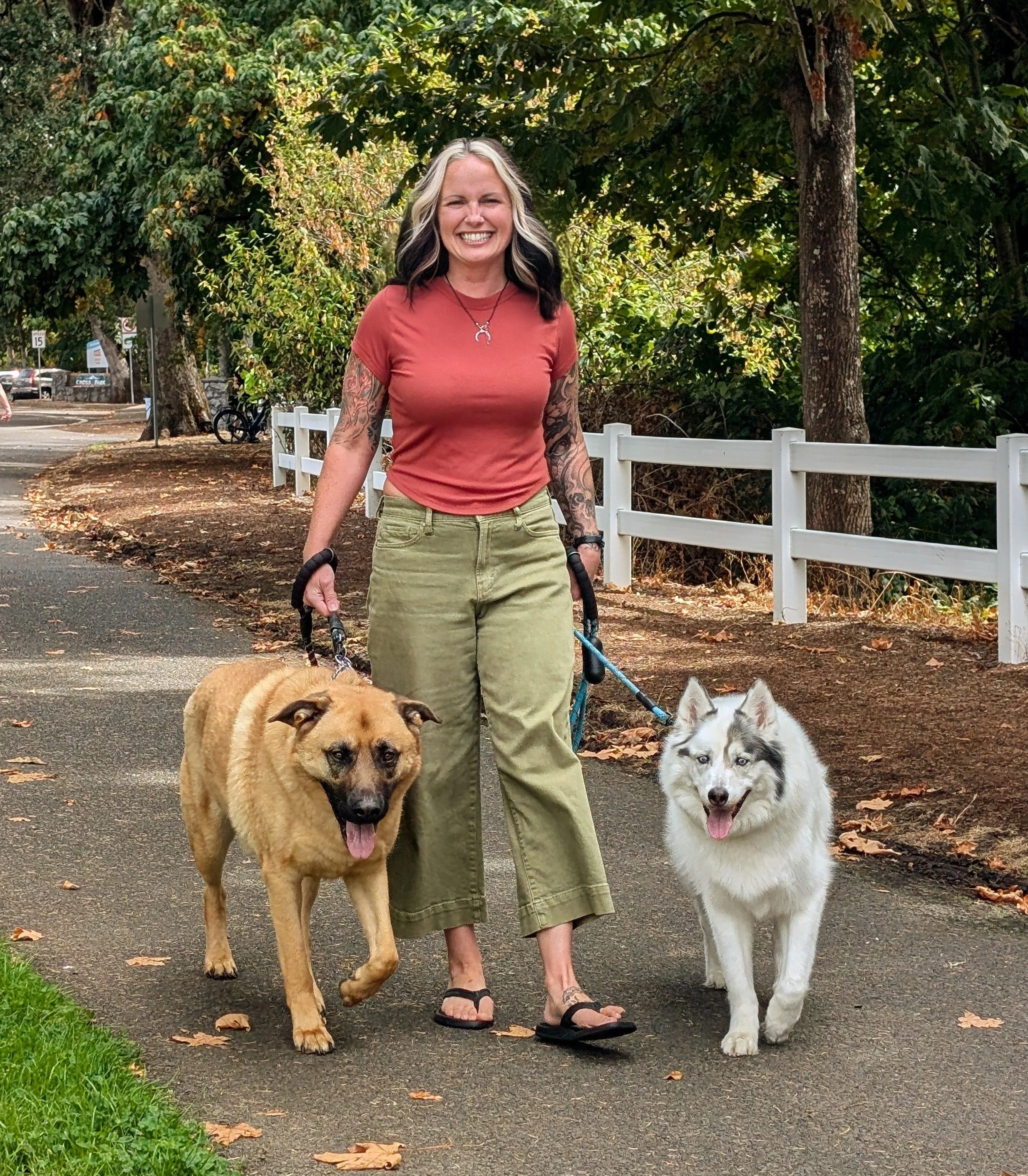 A woman smiling and walking two dogs on a paved path in a park, with trees and a white fence in the background.