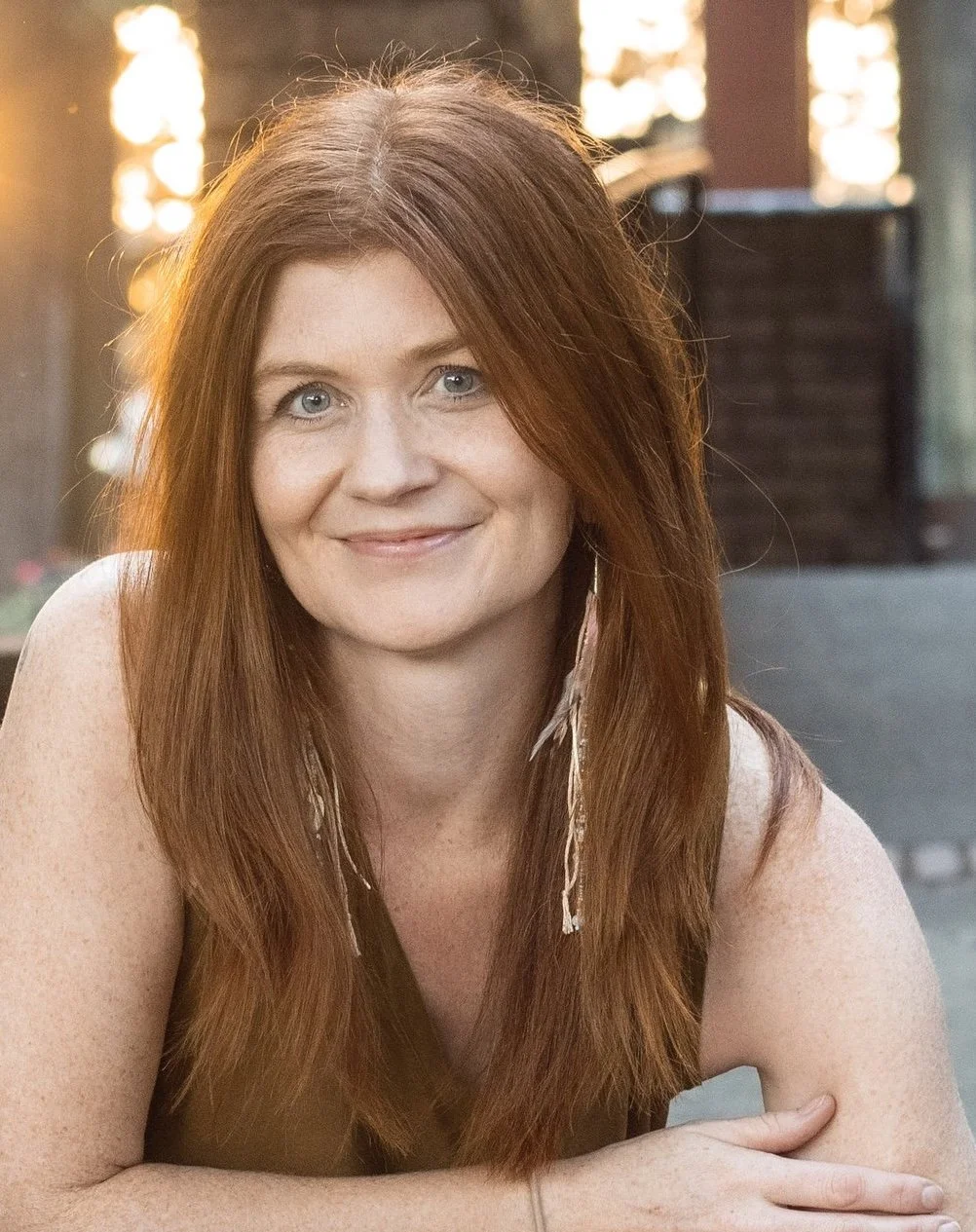 A woman with long red hair and blue eyes smiling, outdoors with warm sunlight and bokeh background.