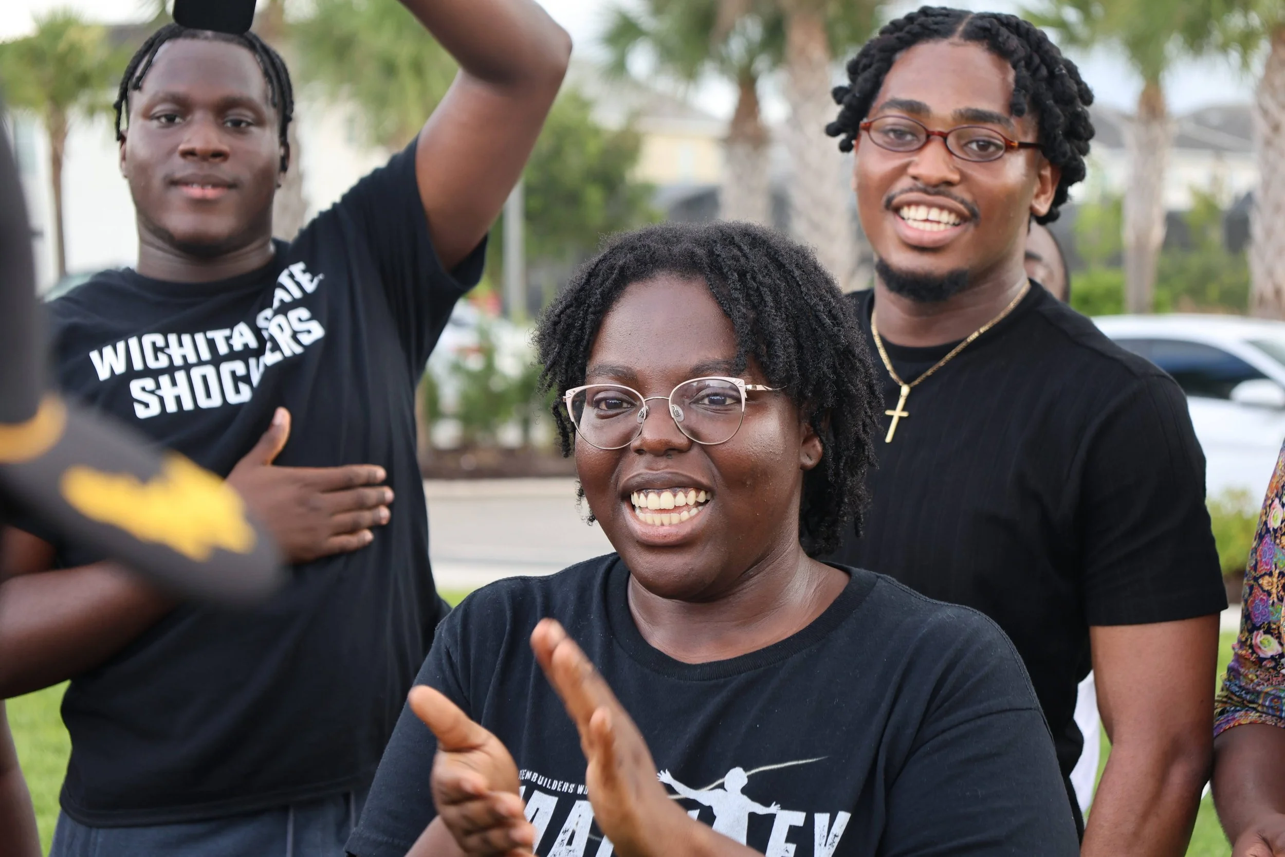 A group of diverse people outdoors, smiling and engaging with each other, with trees and parked cars in the background.