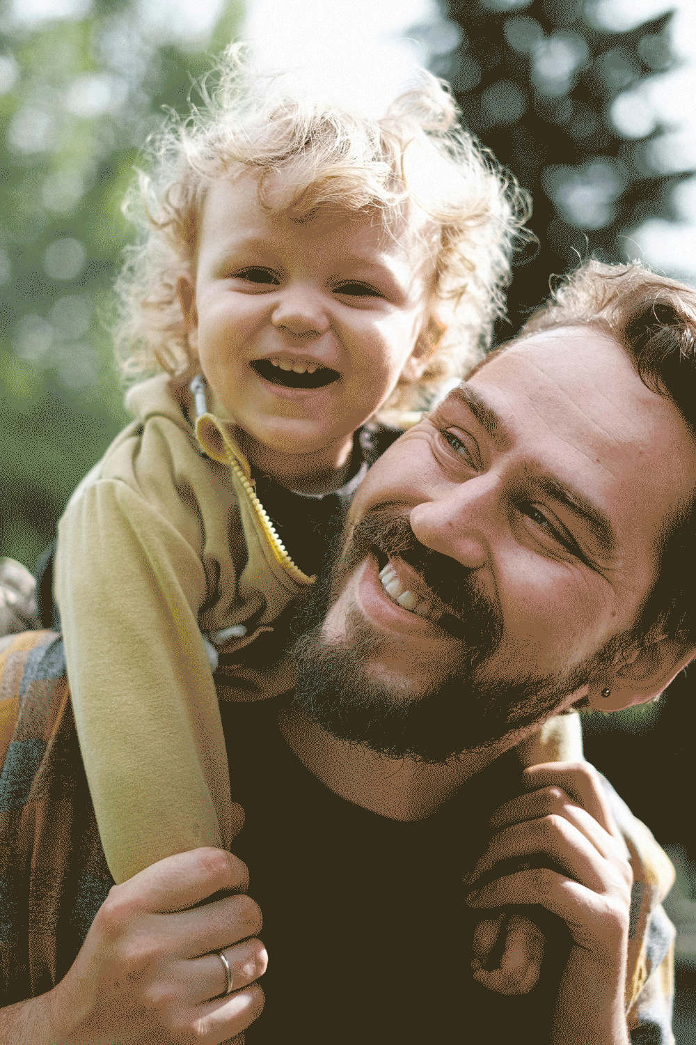 A smiling man with a beard and a child with curly blonde hair, both outdoors on a sunny day.