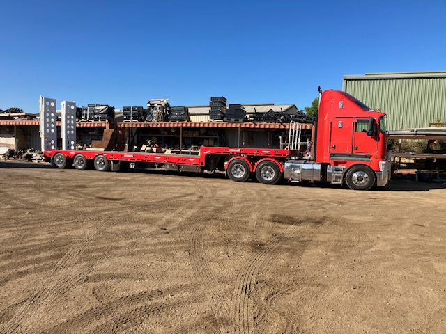 Red flatbed semi-truck parked on a dirt lot with industrial buildings in the background.