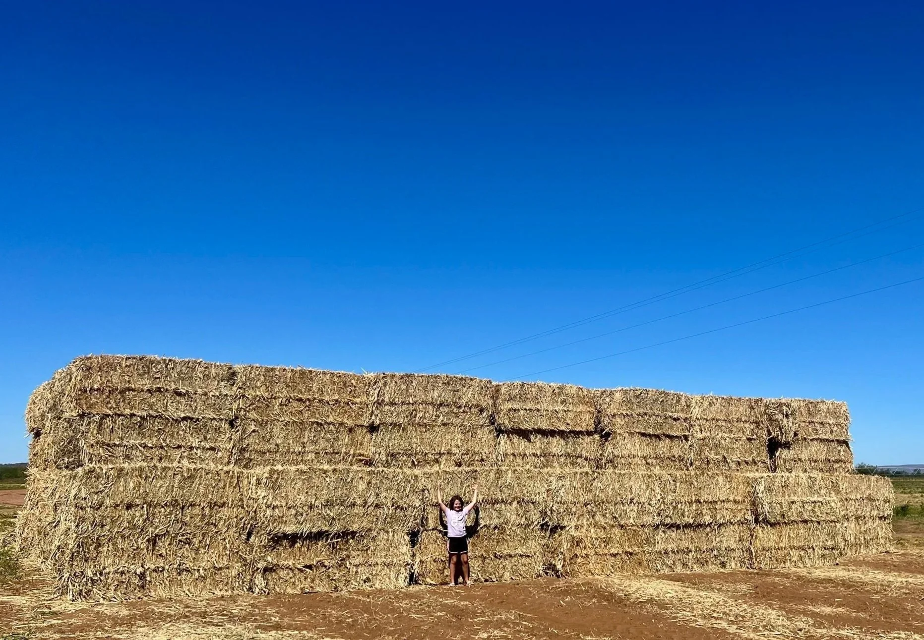 Child standing in front of a large stack of hay bales outdoors under a clear blue sky.