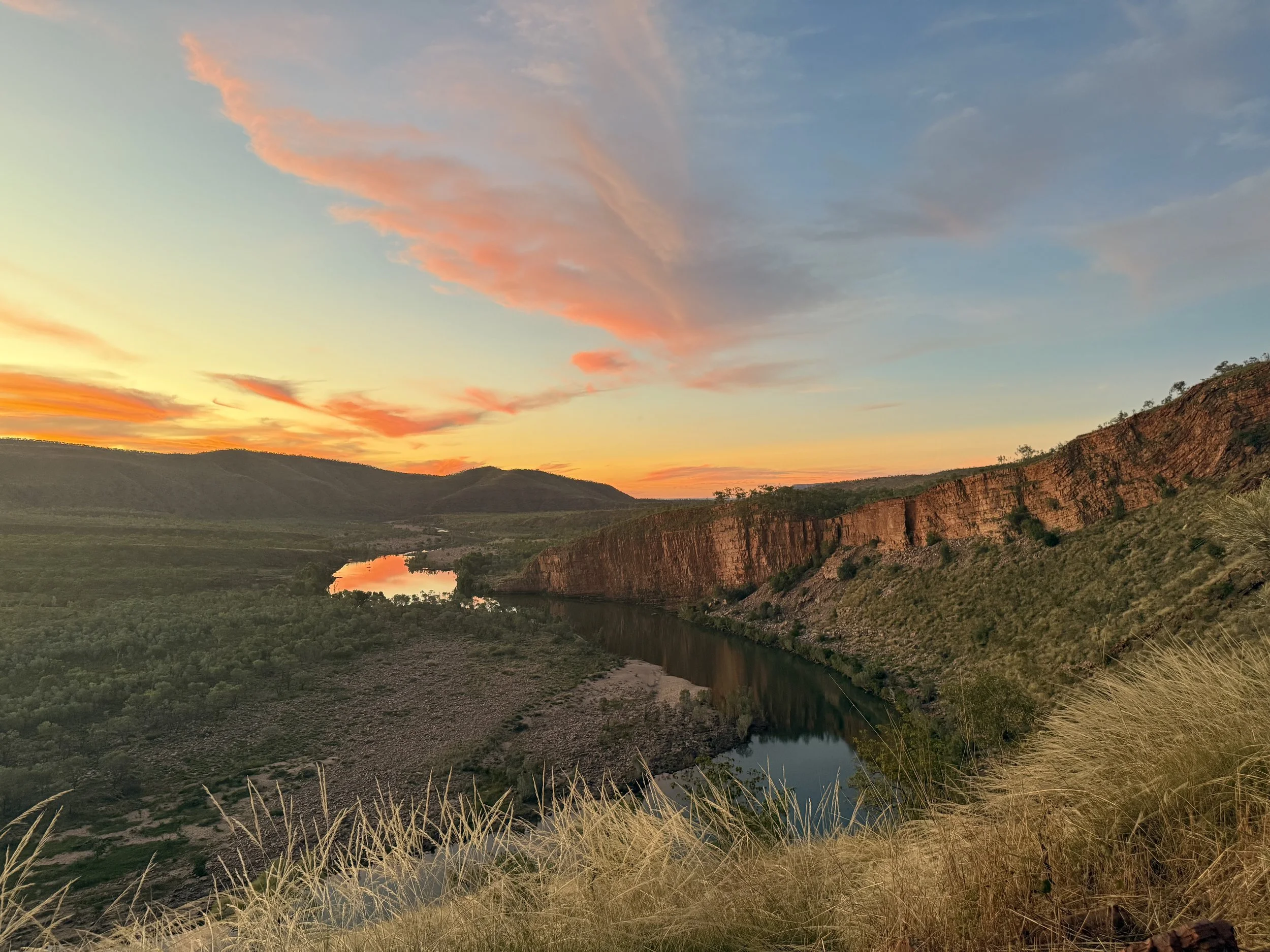 Scenic view of a river flowing through a canyon with green hills, and a colorful sky at sunset with pink and orange clouds.