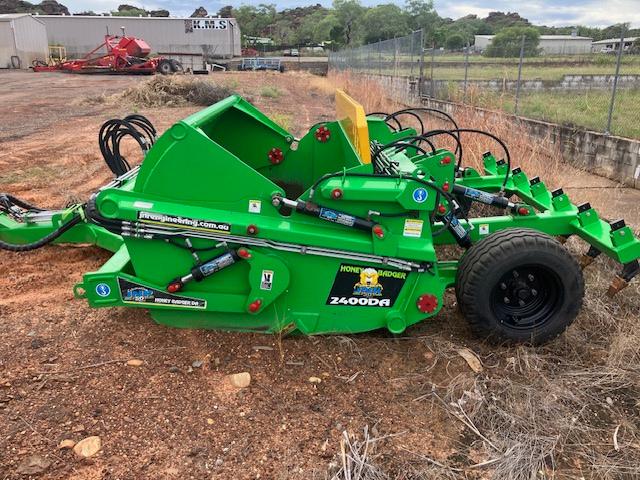 A green agricultural seed drill attached to a tractor, resting on dirt ground in an outdoor setting.