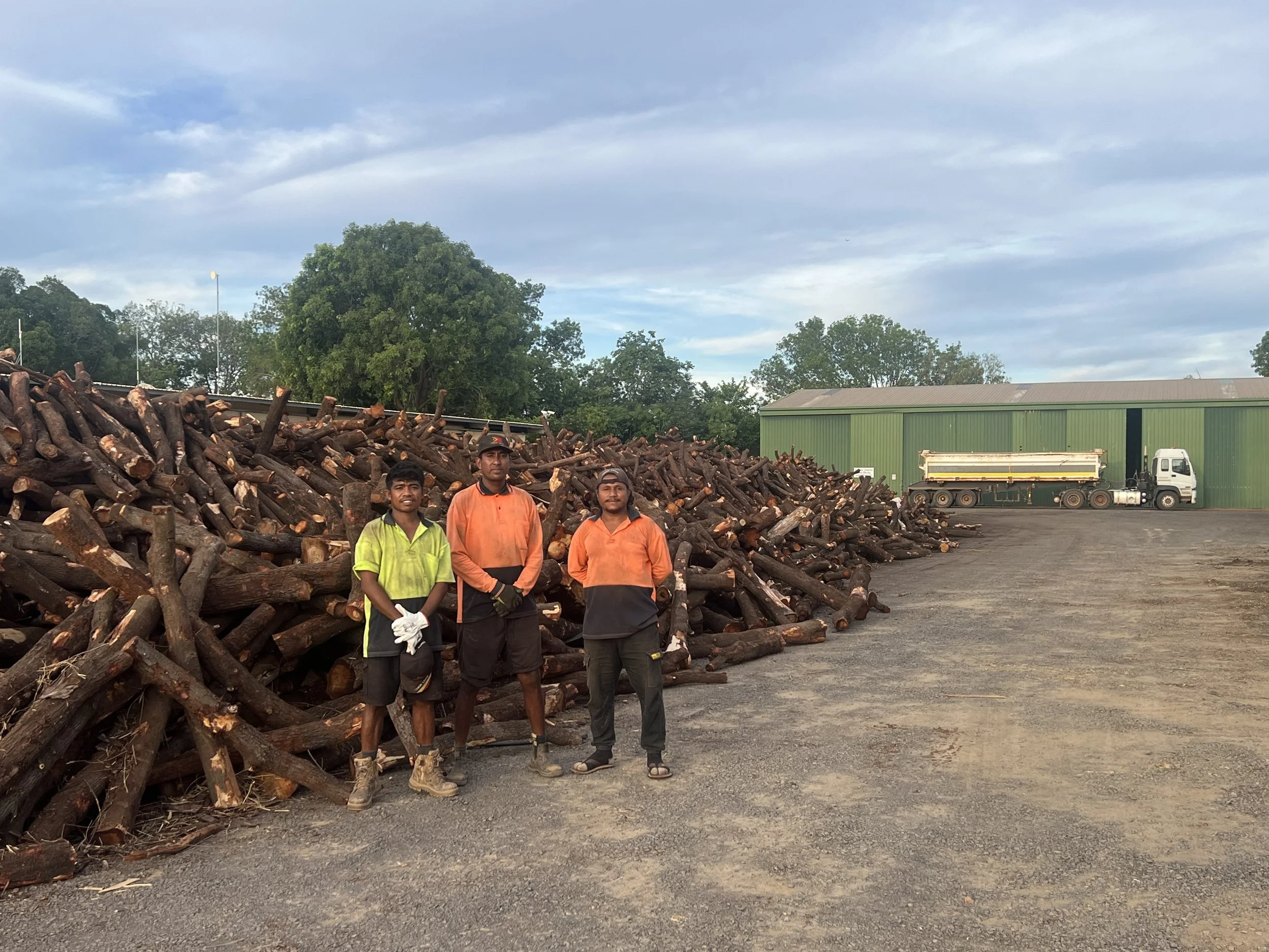 Four men standing in front of a large pile of cut logs. There is a green industrial building and a white truck in the background, with a cloudy sky overhead.