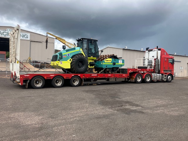 Red semi-truck with a flatbed trailer carrying a large green and yellow construction roller, parked outside in front of industrial buildings under dark cloudy skies.