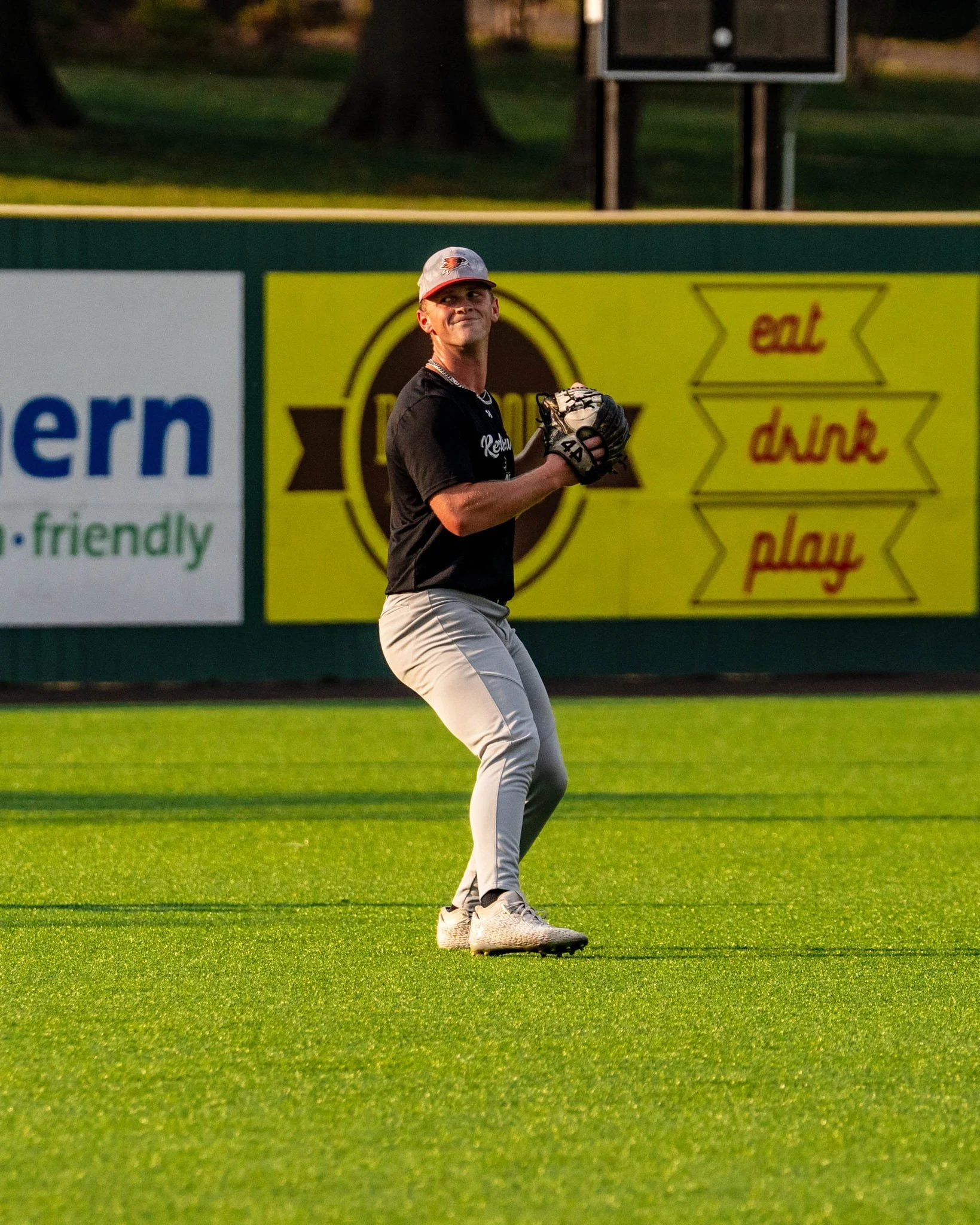 A young man in a black t-shirt and grey baseball pants prepares to catch a baseball on a grassy field during sunset; he's wearing a baseball glove and cap and smiling.