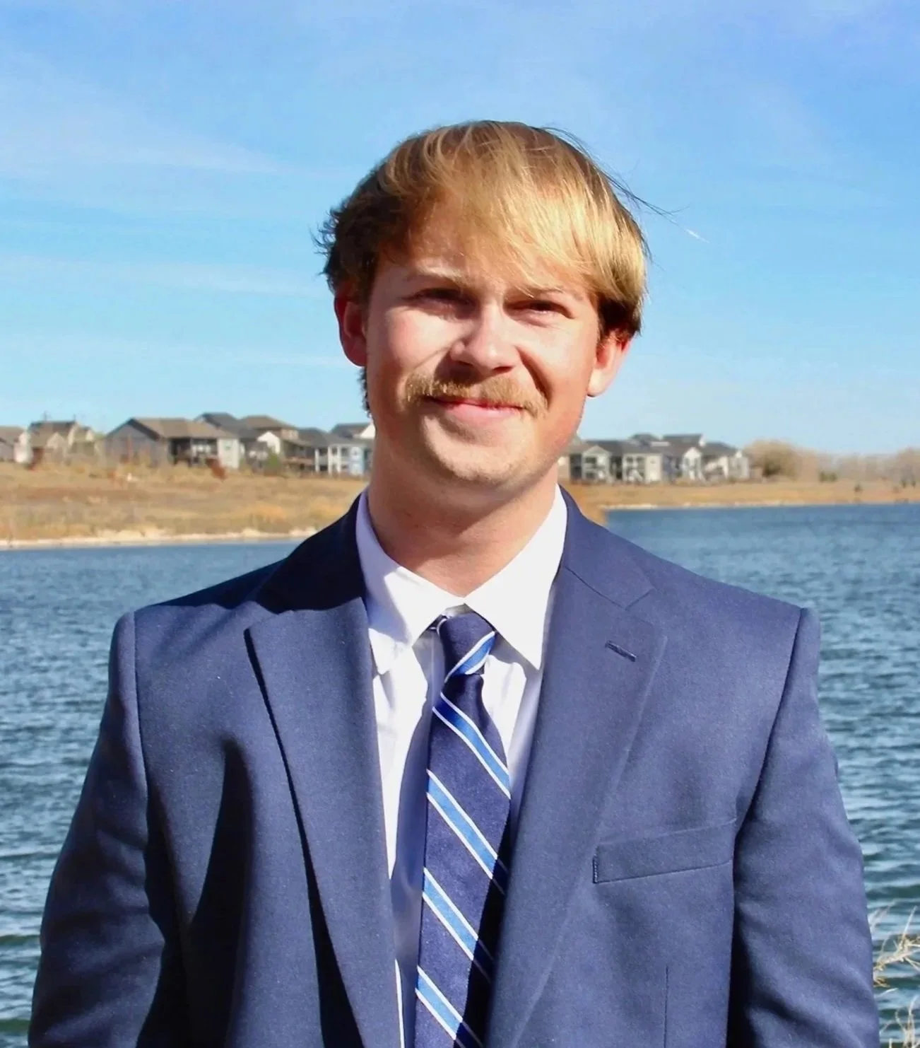 A young man with red hair and a mustache wearing a blue suit, striped tie, and white shirt standing outdoors near a body of water with houses in the background.