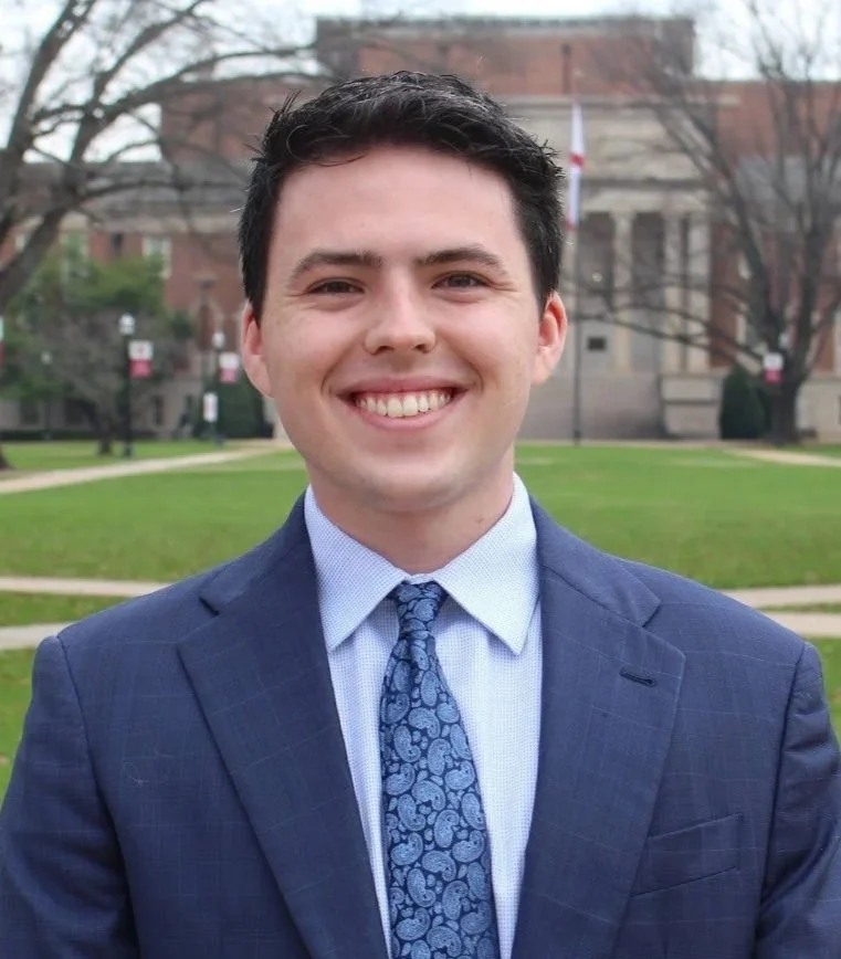 A young man in a blue suit and patterned tie smiling outdoors with a grass lawn, trees, and a historic brick building in the background.