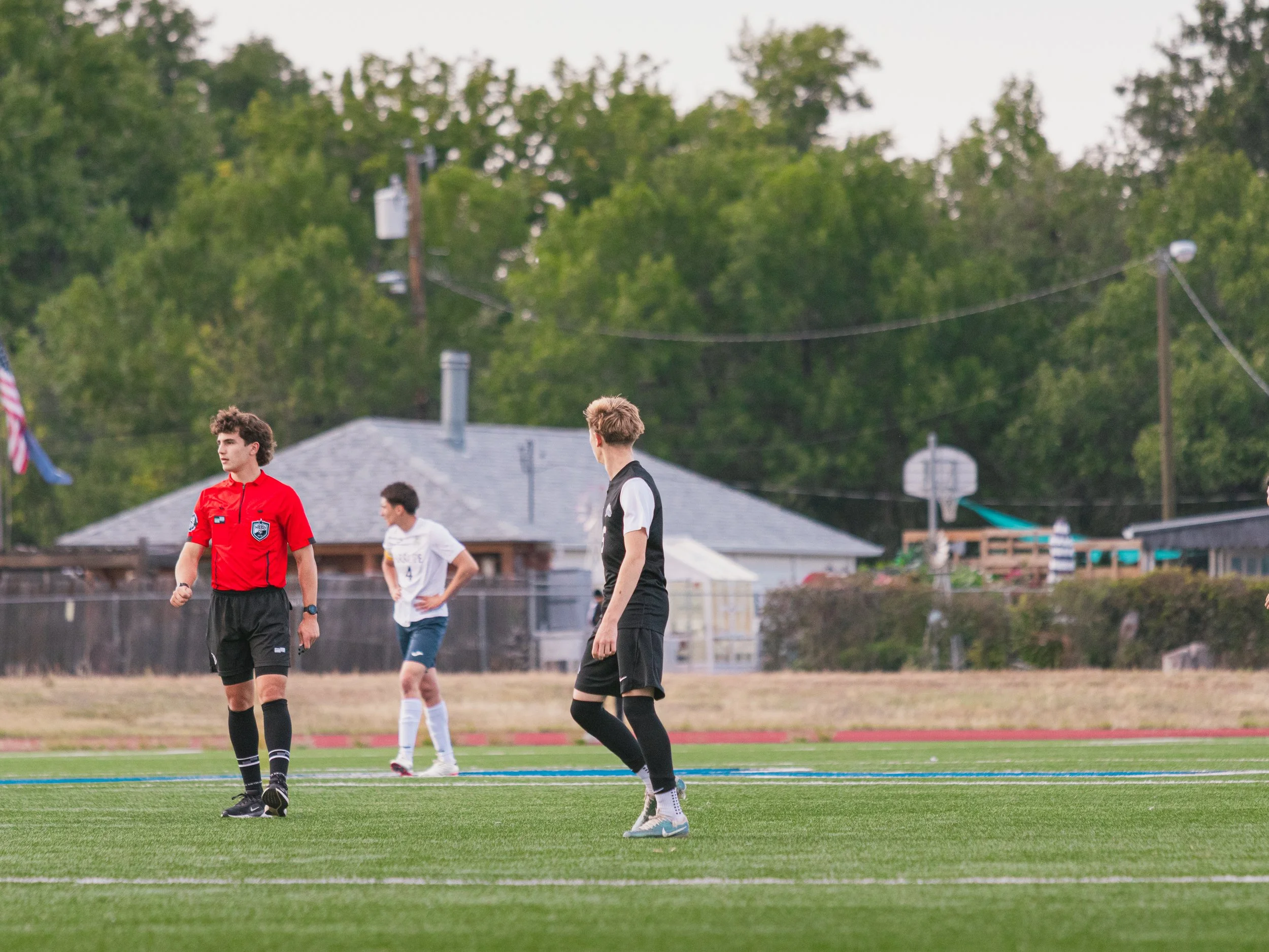 Youth soccer players stand on a field with a referee in a red shirt, with a house and trees in the background.