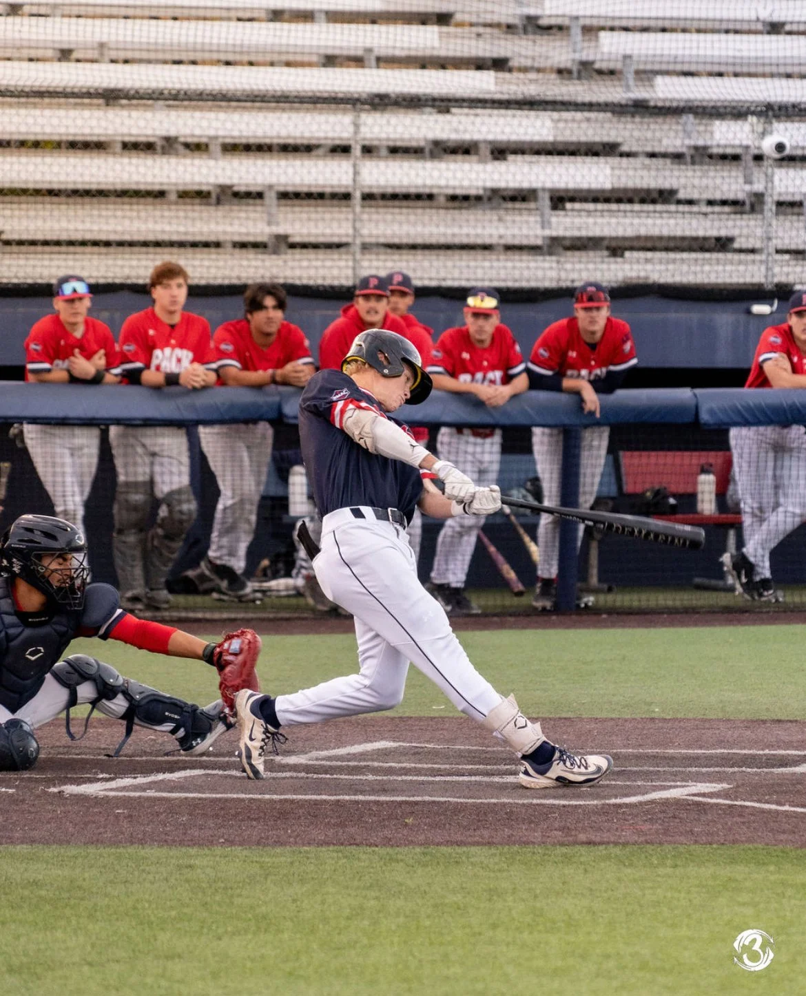 A baseball player in a navy and white uniform swinging a bat at a pitch on the field, with team members watching from the dugout in the background.