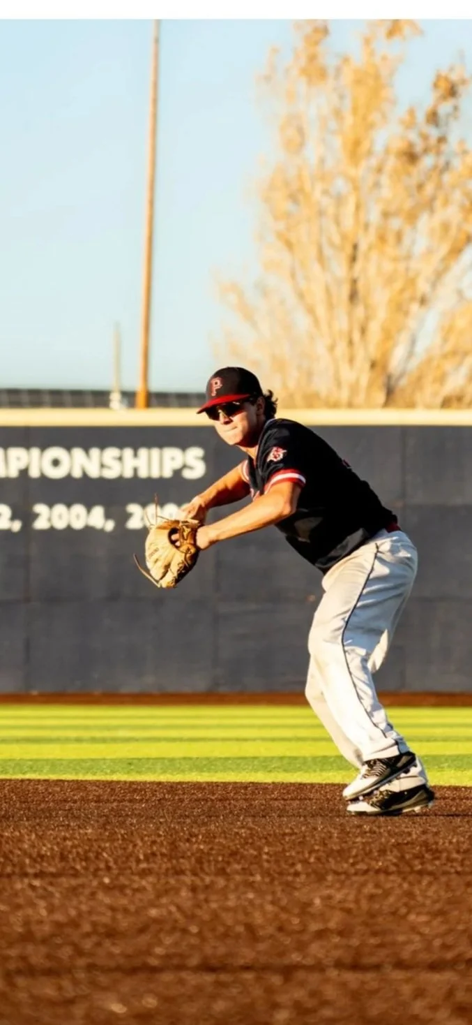 A baseball player in a black uniform with a red cap and sunglasses fielding a ball on a baseball field during daytime.