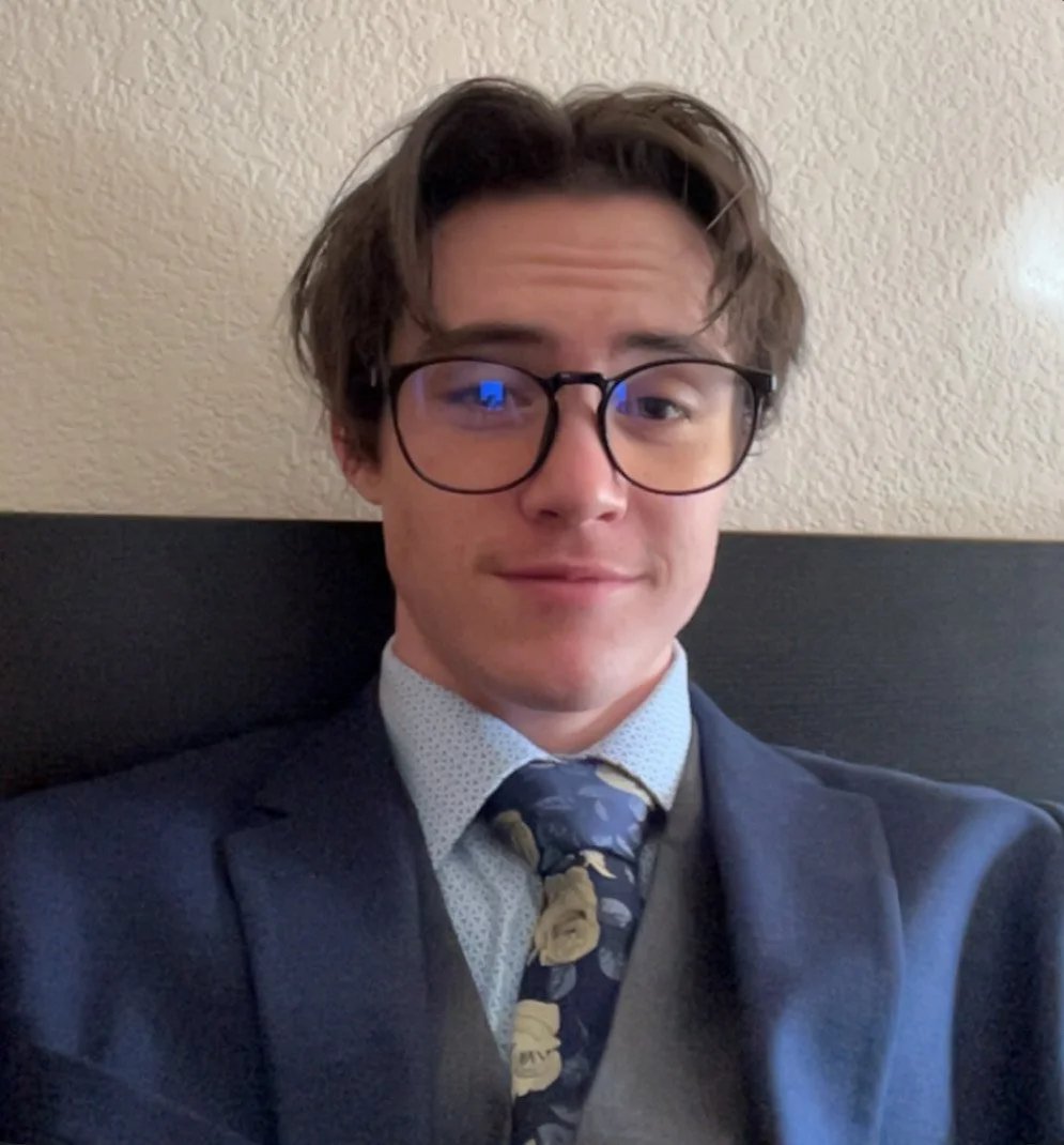 Young man with glasses in a suit and tie taking a selfie indoors against a beige textured wall.