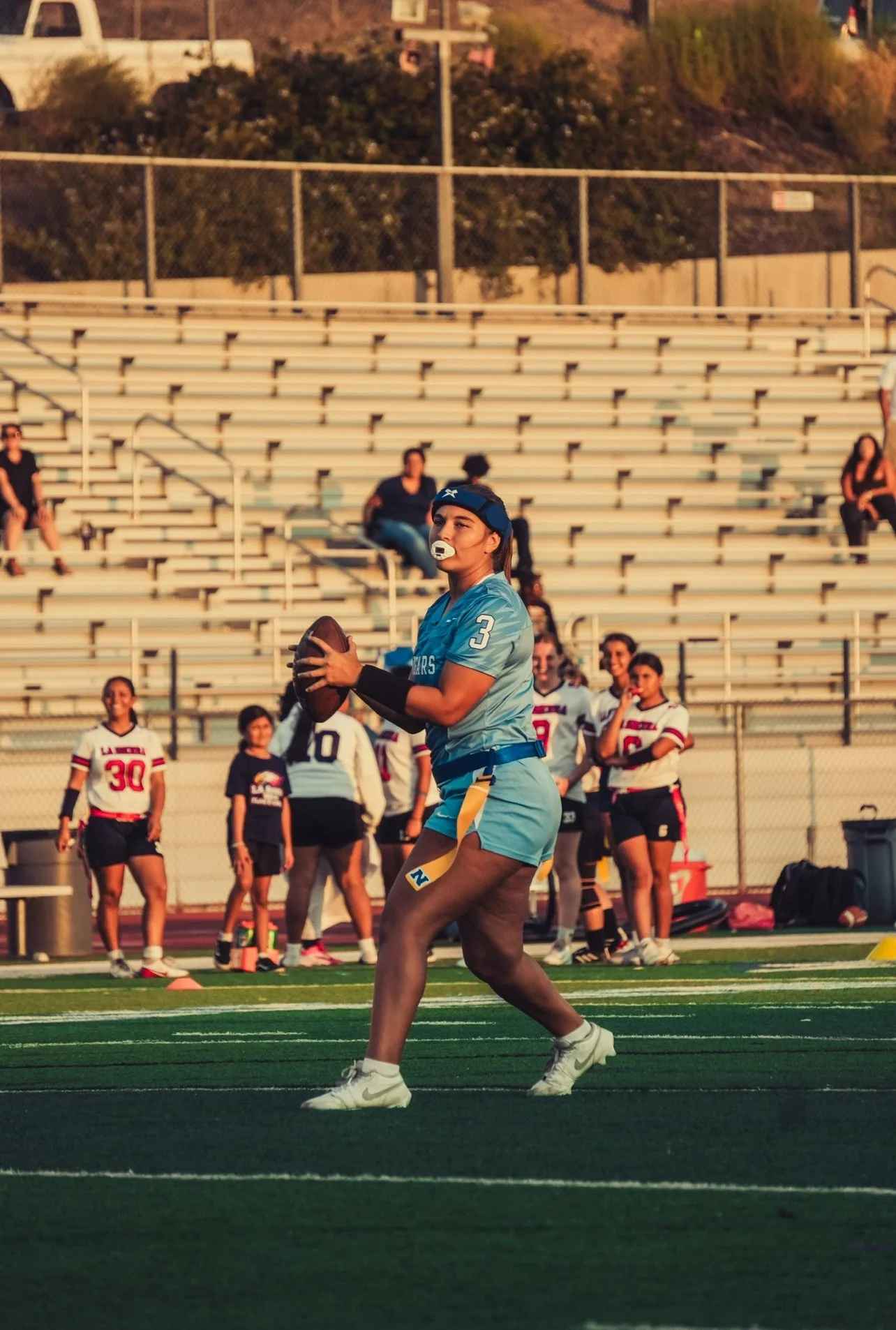 A female flag football player in a blue uniform with the number 3, holding a football, on a field during a game. In the background, other players and spectators are present, with empty bleachers and a chain-link fence.