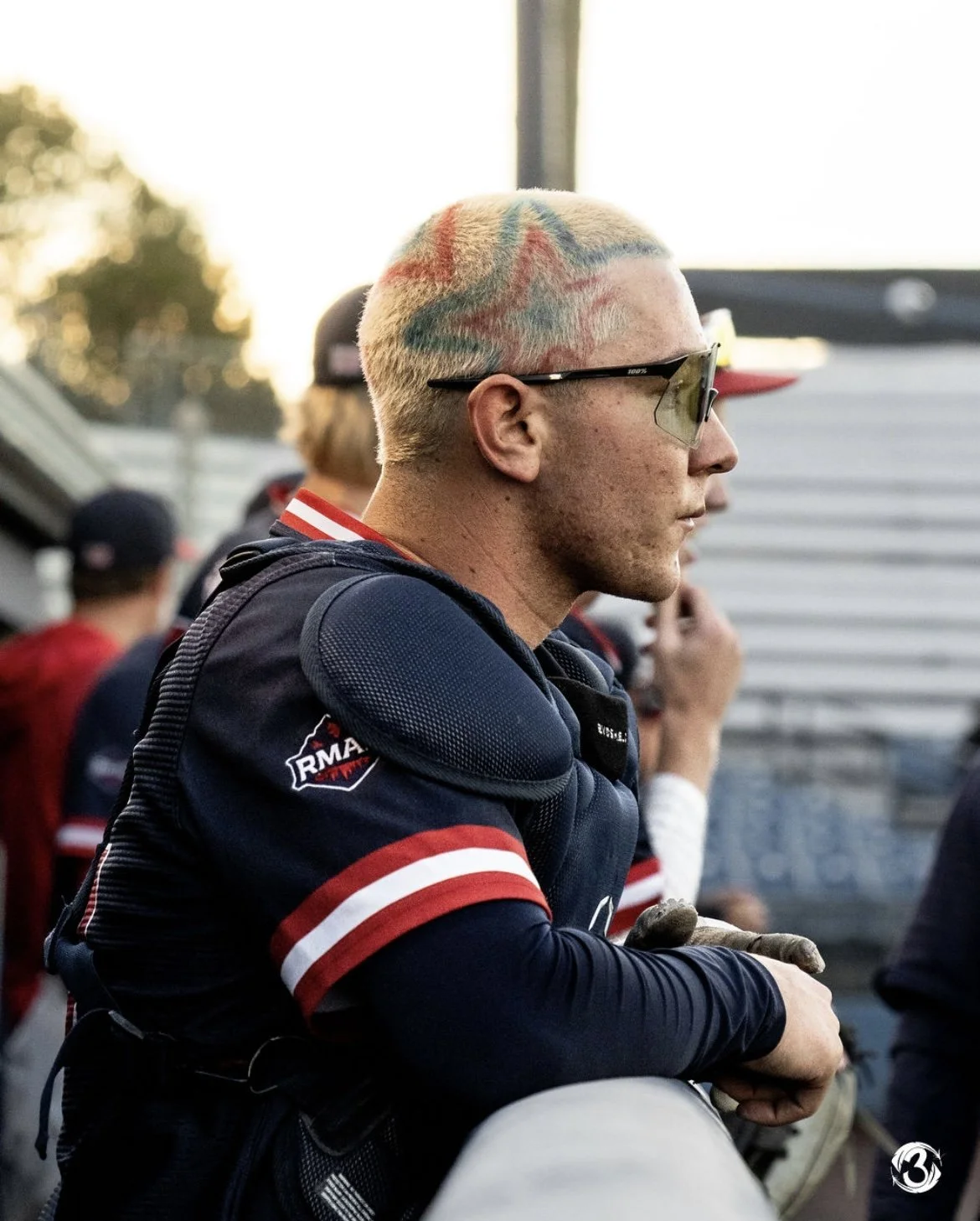 A man with dyed rainbow-colored hair wearing sunglasses and a racing suit, sitting with arms crossed at a racetrack paddock.