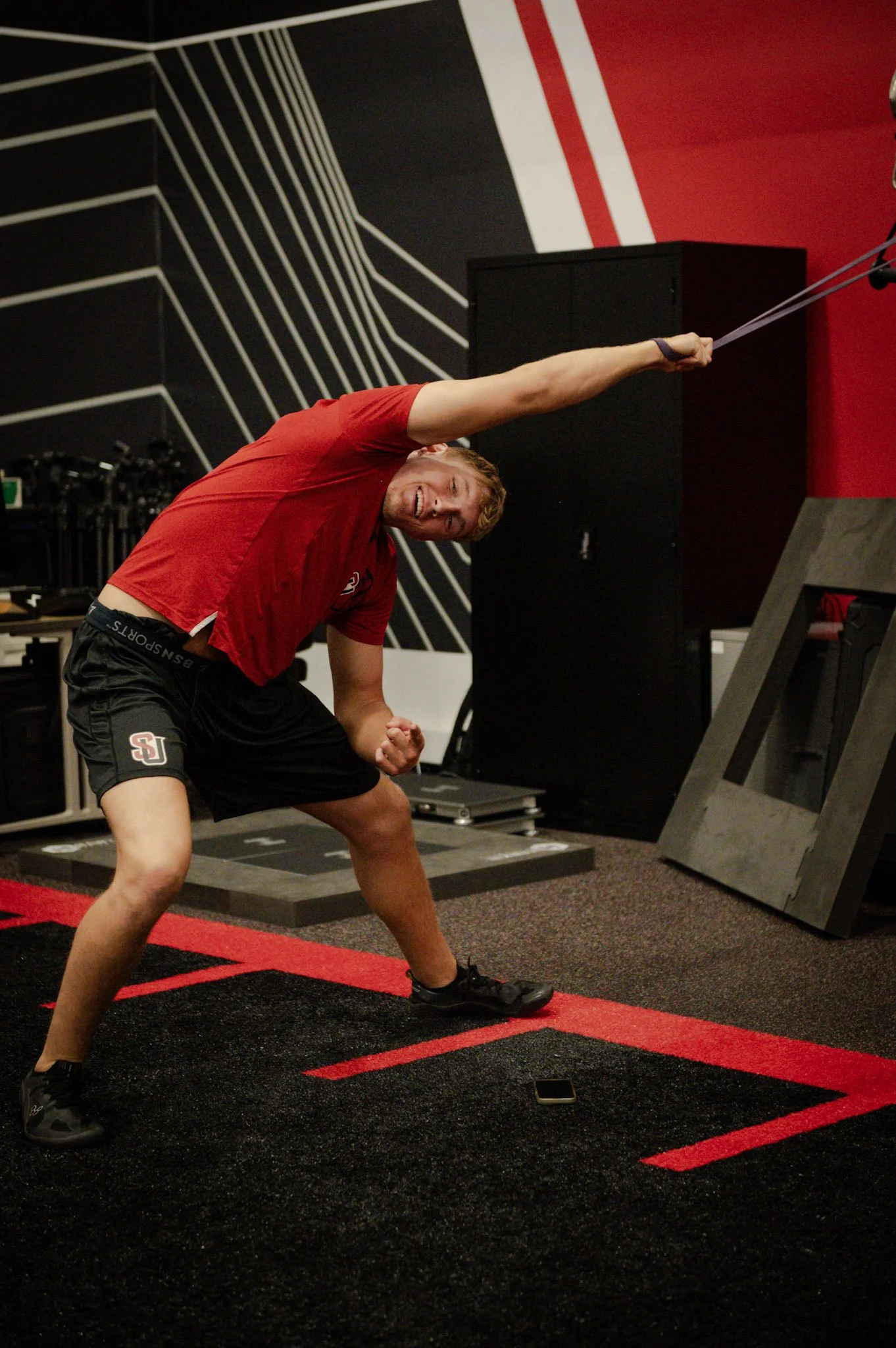 Travis Farrell, pitcher formerly at CSU Pueblo and currently at Seattle University. Workout, stretch, athlete, resistance. Young man in red shirt and black shorts exercising in a gym, pulling resistance band overhead with determined expression.