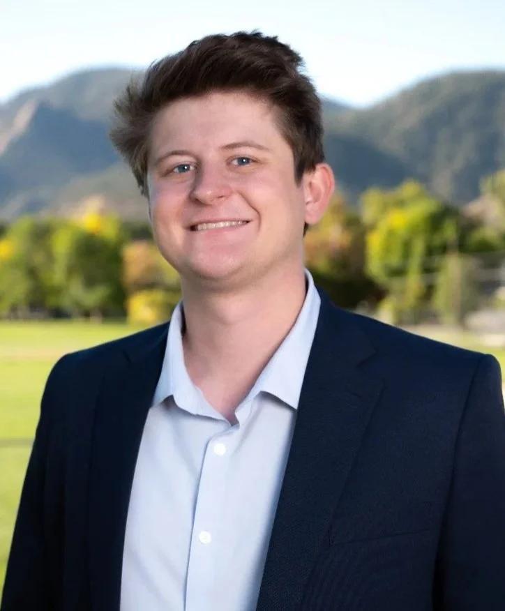A young man in a navy blazer and light blue dress shirt outdoors with mountains and trees in the background.