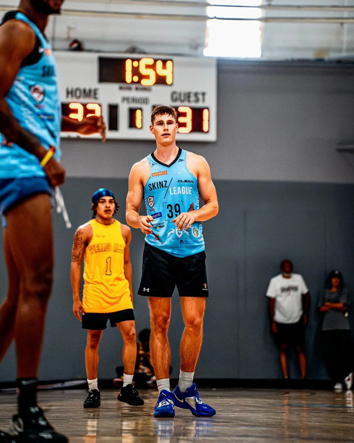 A young male relay athlete in a blue jersey and black shorts standing on an indoor basketball court with a scoreboard in the background showing 1:54 remaining and a score of 33-31.
