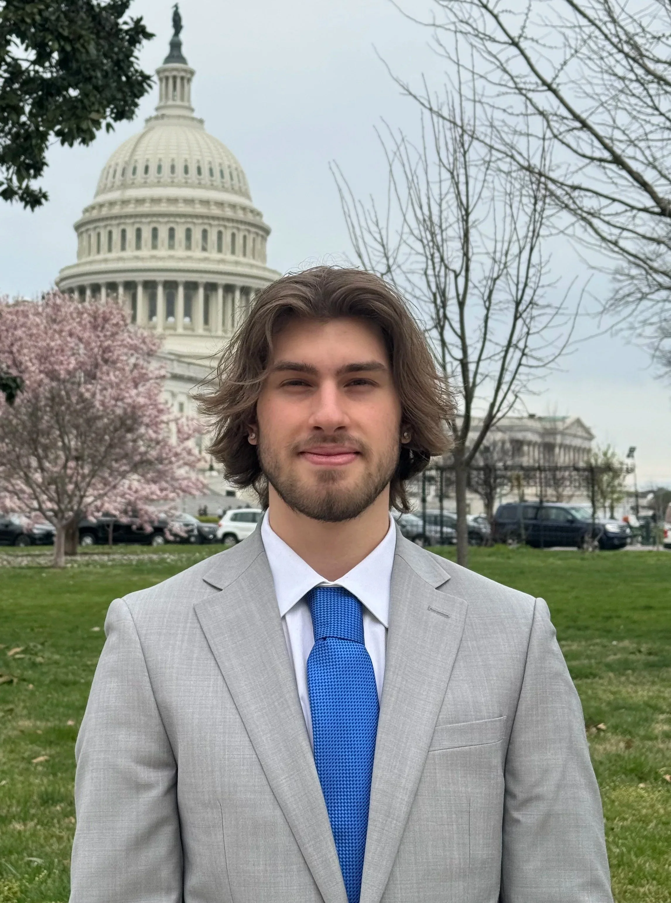 A young man with shoulder-length brown hair and a beard, wearing a light gray suit, white shirt, and blue tie, standing outdoors in front of the U.S. Capitol building with cloudy sky, blooming trees, and parked cars in the background.