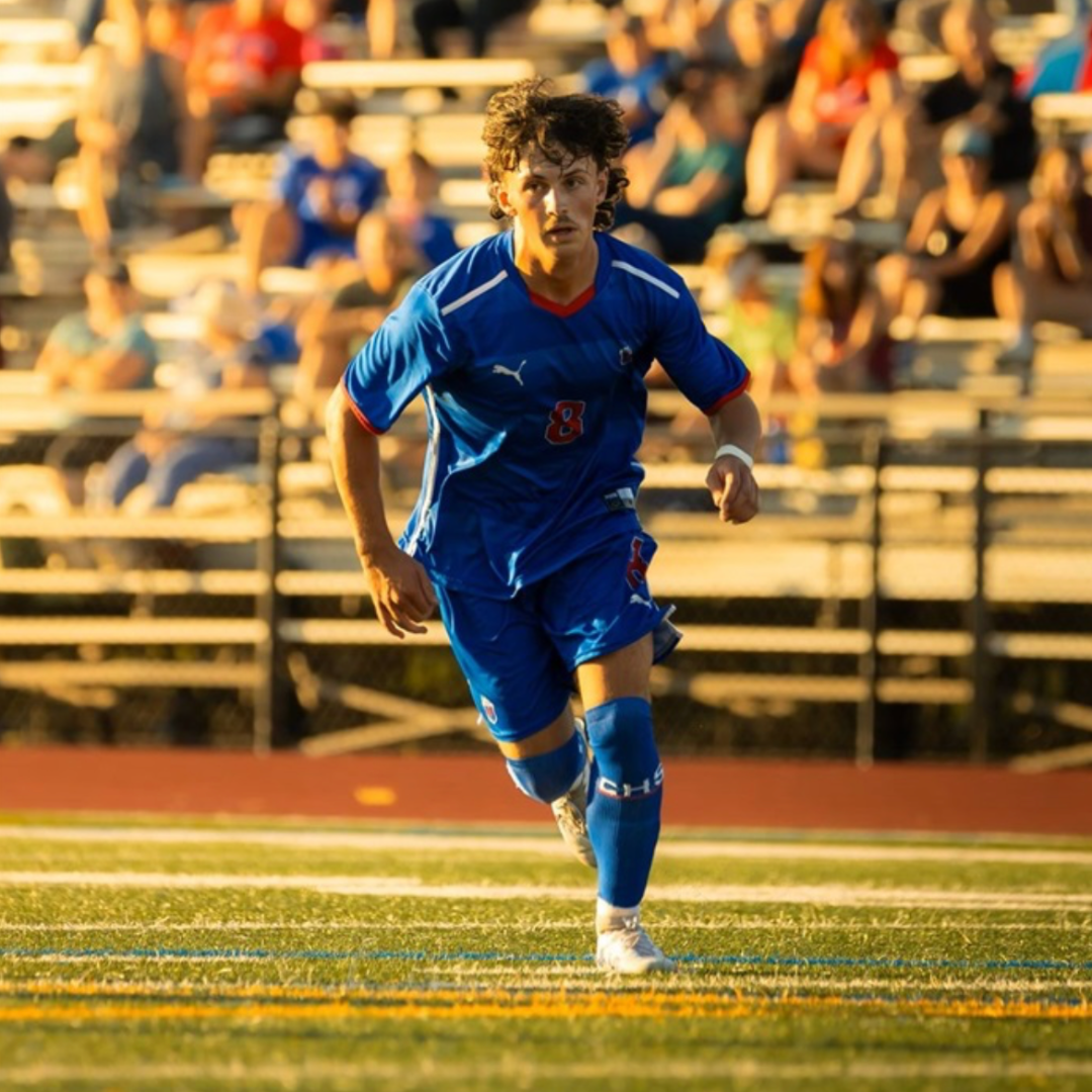 A young male soccer player in a blue uniform running on a field with spectators in the background during a game.