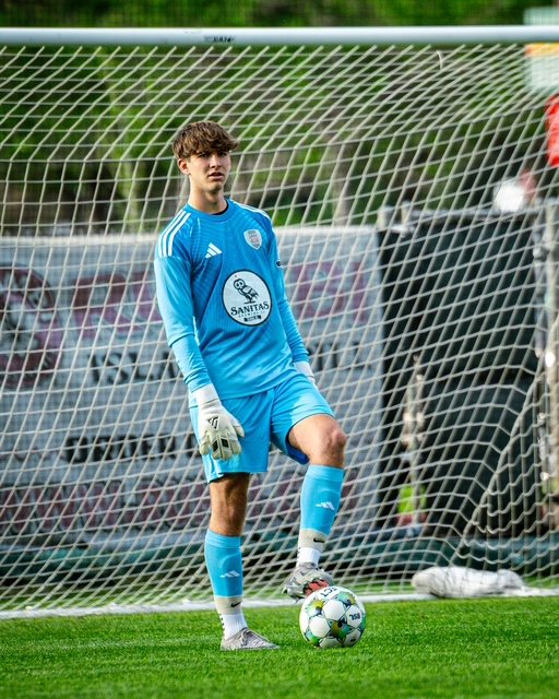 A young soccer goalkeeper standing in front of the goal, wearing a light blue uniform, gloves, and white soccer cleats, with a soccer ball on the ground.