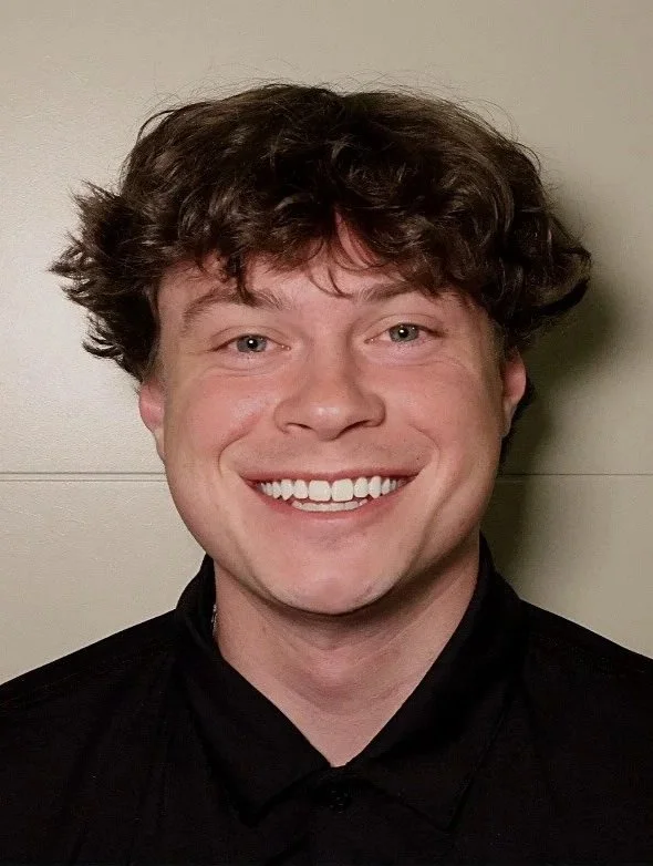 A young man with curly brown hair, fair skin, and blue eyes, smiling at the camera, wearing a black shirt, standing against a light-colored wall.