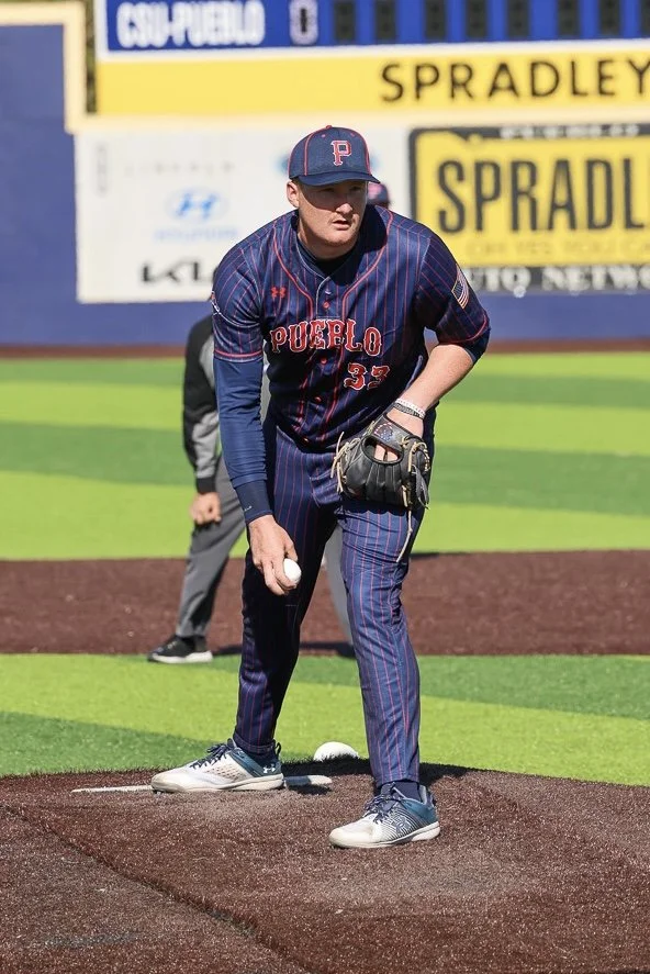 A baseball player in a navy blue pinstripe uniform getting ready to pitch on the mound. The player is wearing a cap with a P on it, and the jersey has 'PUEBLO' and the number 33. There is a person in the background standing on the field and a yellow advertising banner on the outfield wall.