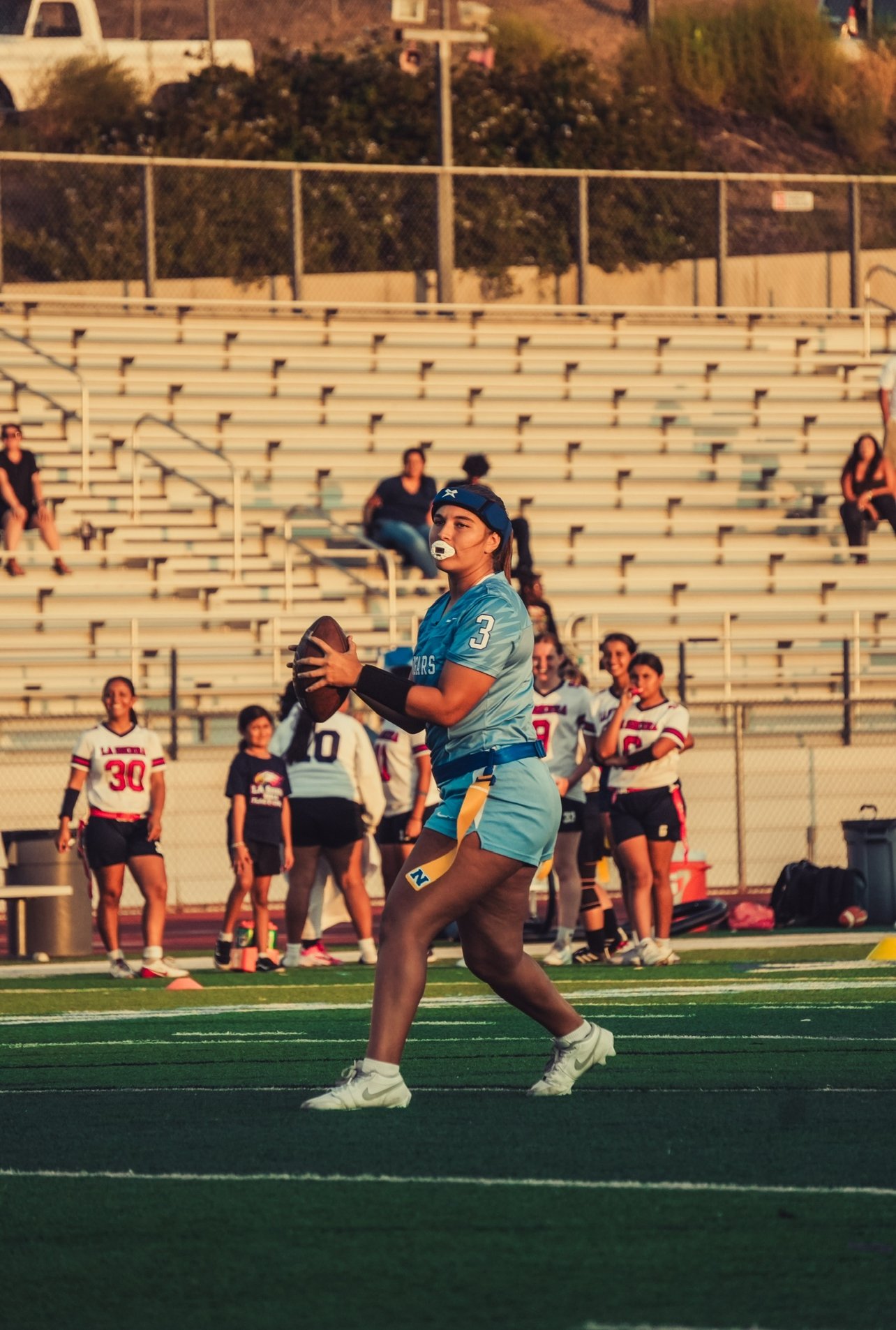 A female football player in a blue uniform holding a football on a field with other players and spectators in the background.