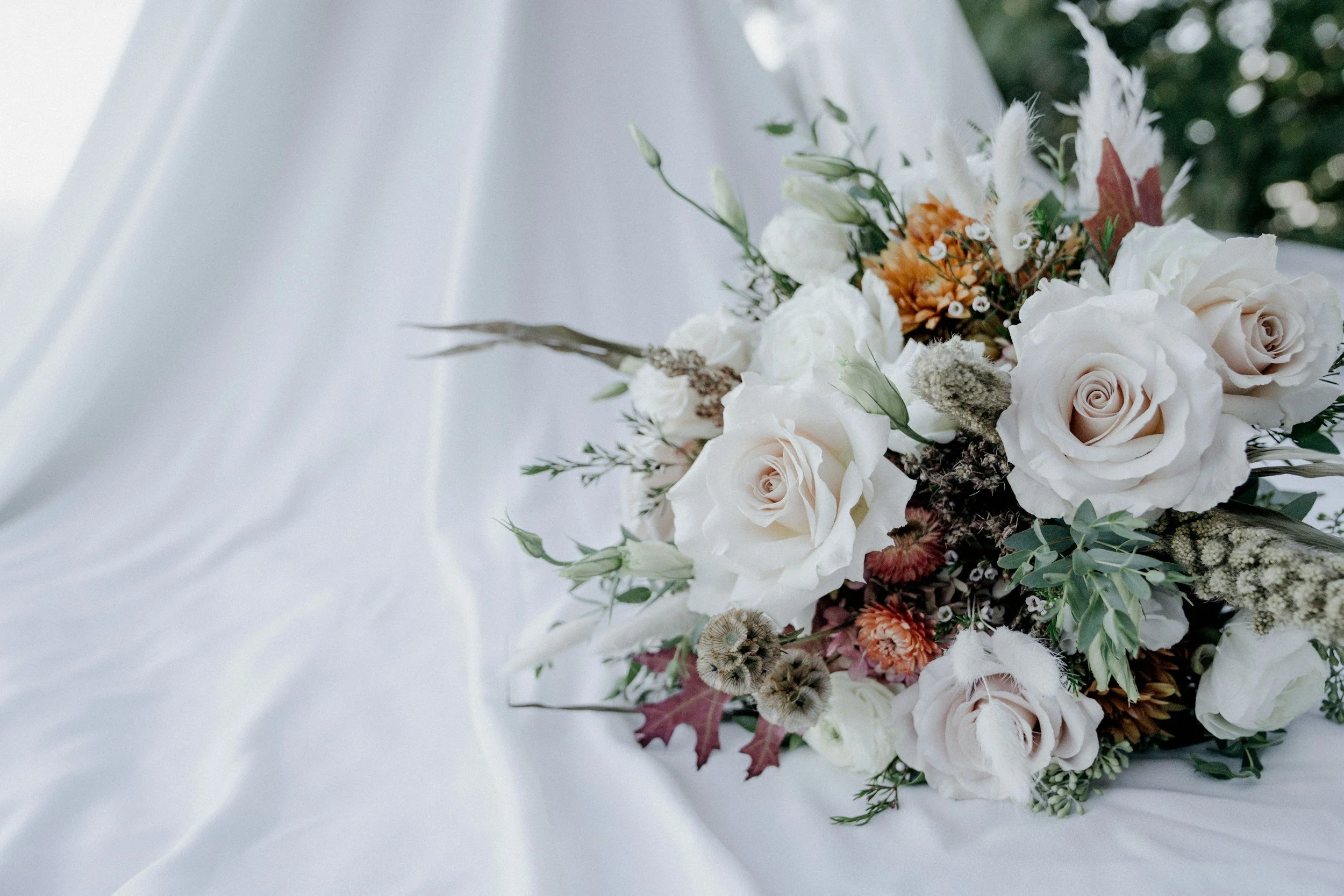 A bouquet of white and light pink roses with greenery and other flowers on white fabric.