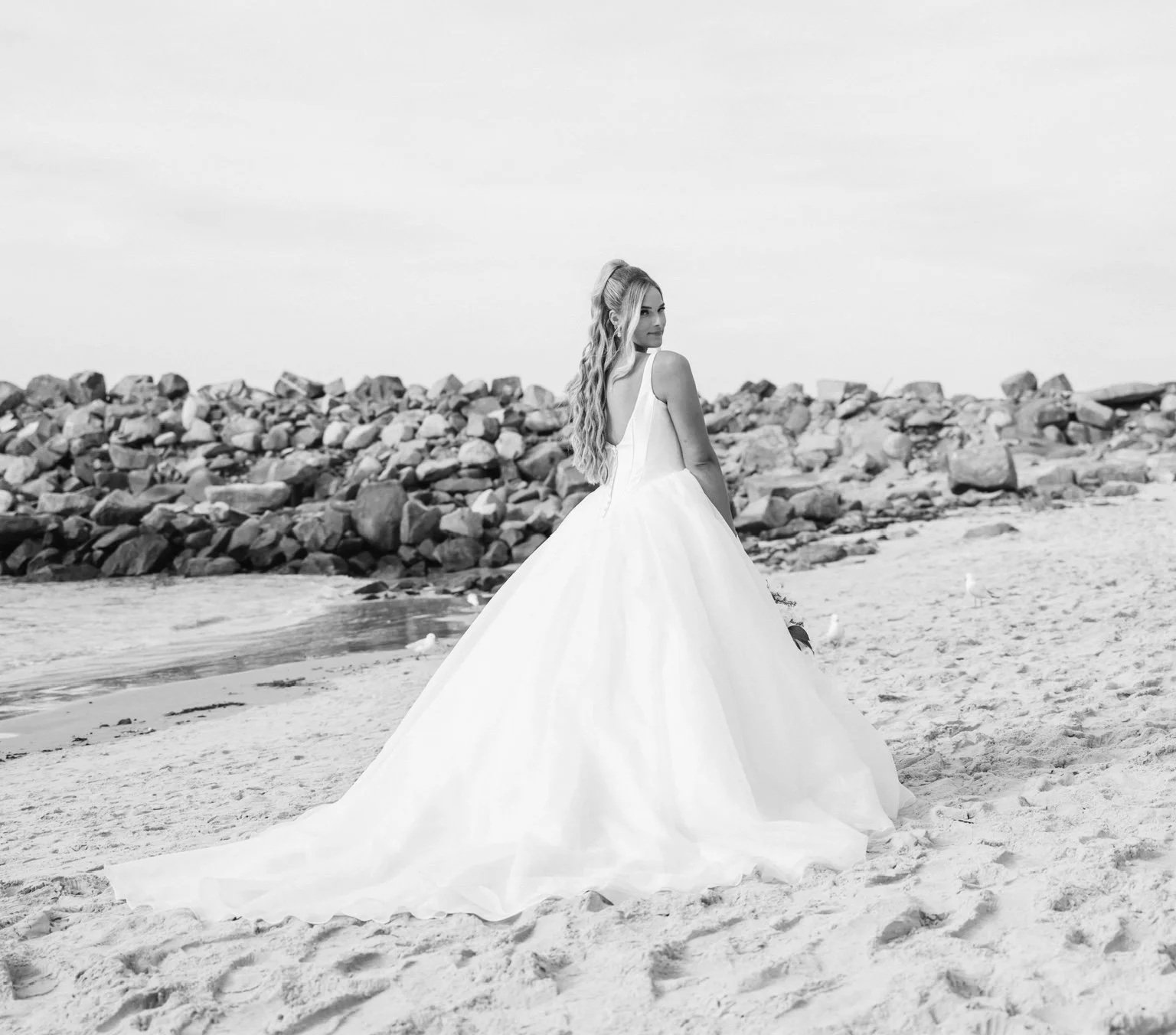 A woman in a wedding dress standing on a sandy beach, looking over her shoulder with a stone barrier in the background.