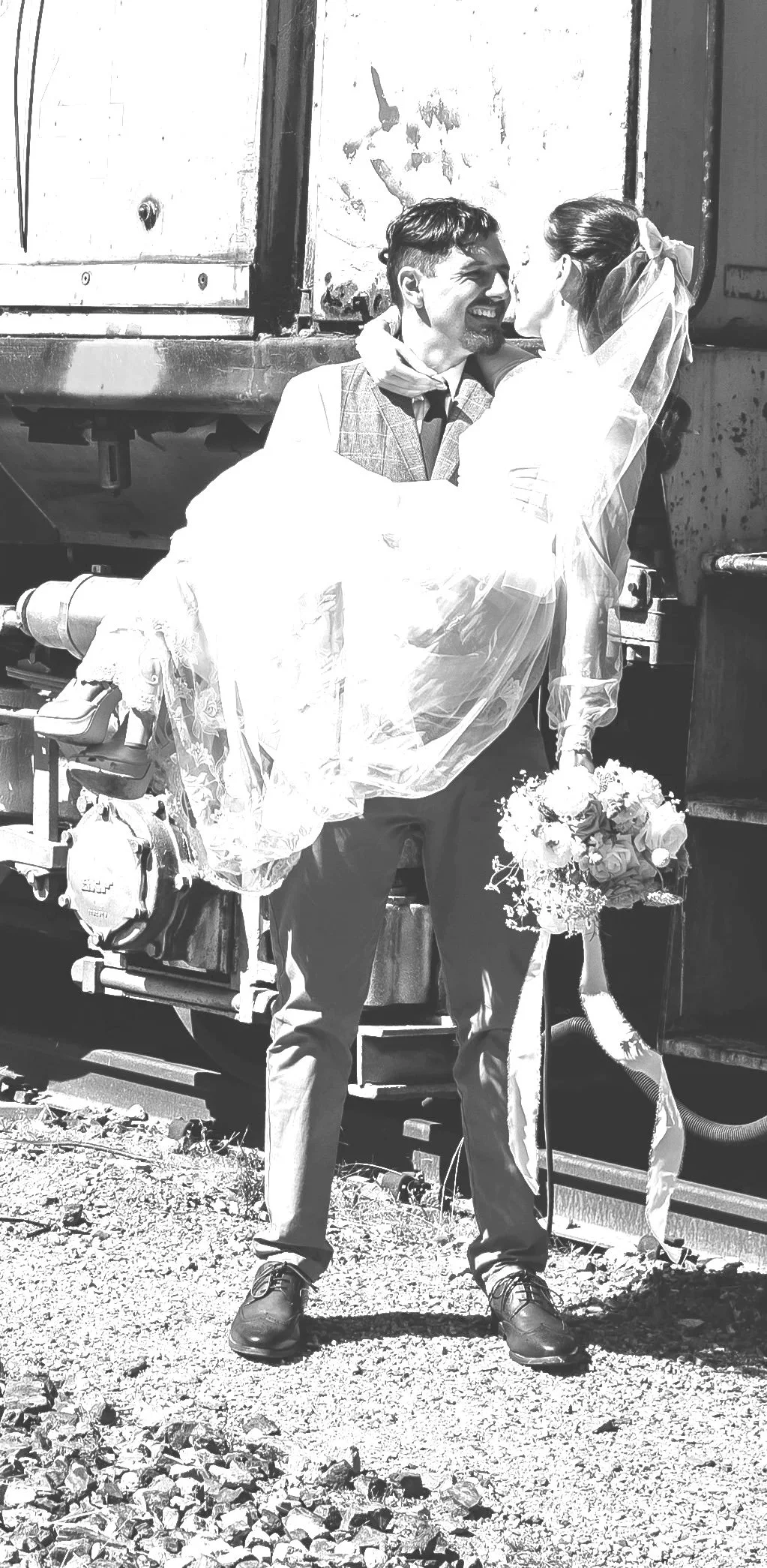 Black and white photo of a man holding a woman in a bridal dress. The woman holds a wedding bouquet, and they are smiling at each other. They are standing outdoors in front of a weathered train car.