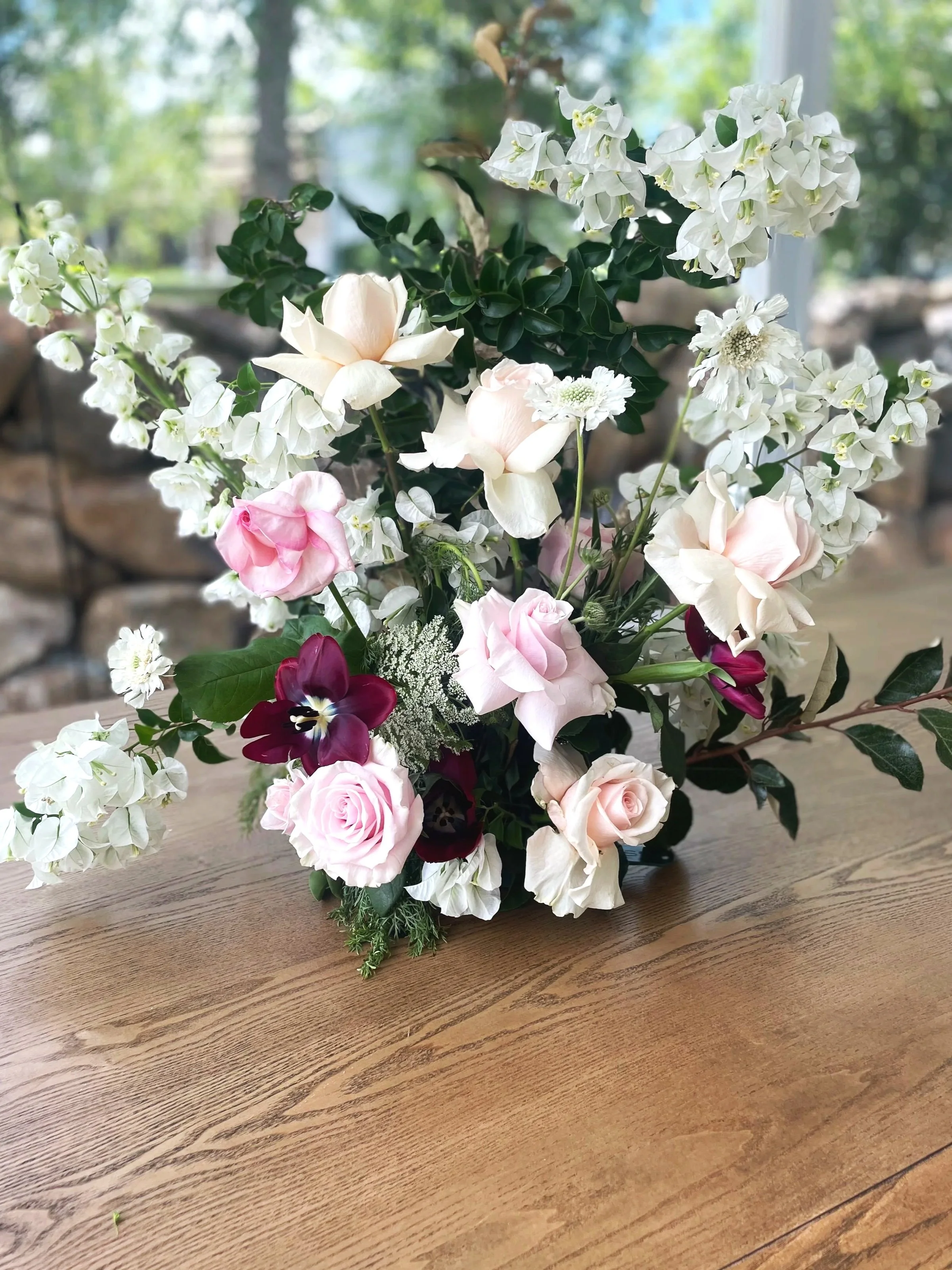 A wedding floral arrangement with white, pink, and dark red flowers, including roses, pansies, and other blossoms, on a wooden table with a blurred outdoor background.