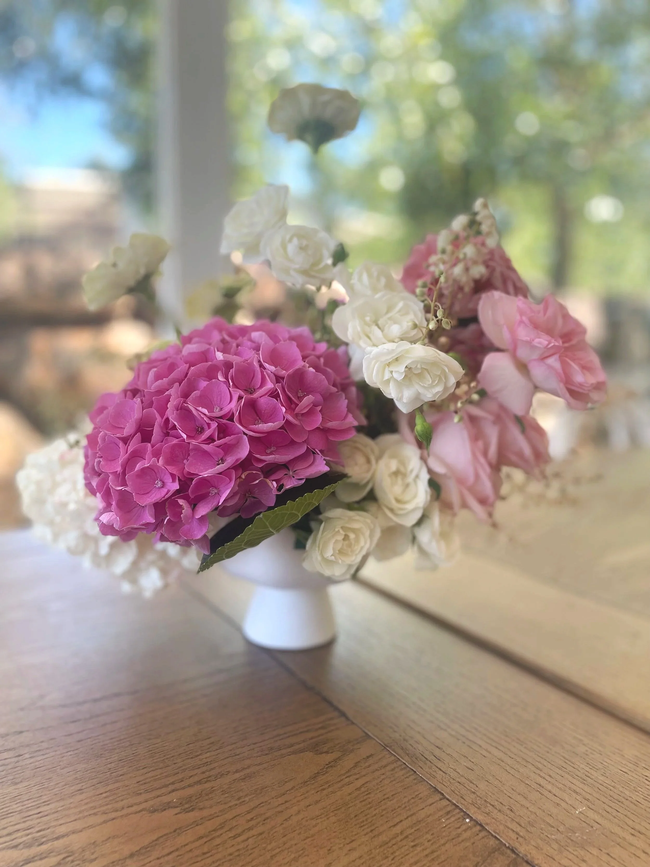 A wedding floral arrangement with pink, white, and light pink flowers in a small white vase on a wooden table, with a blurred background showing a window and greenery outside.