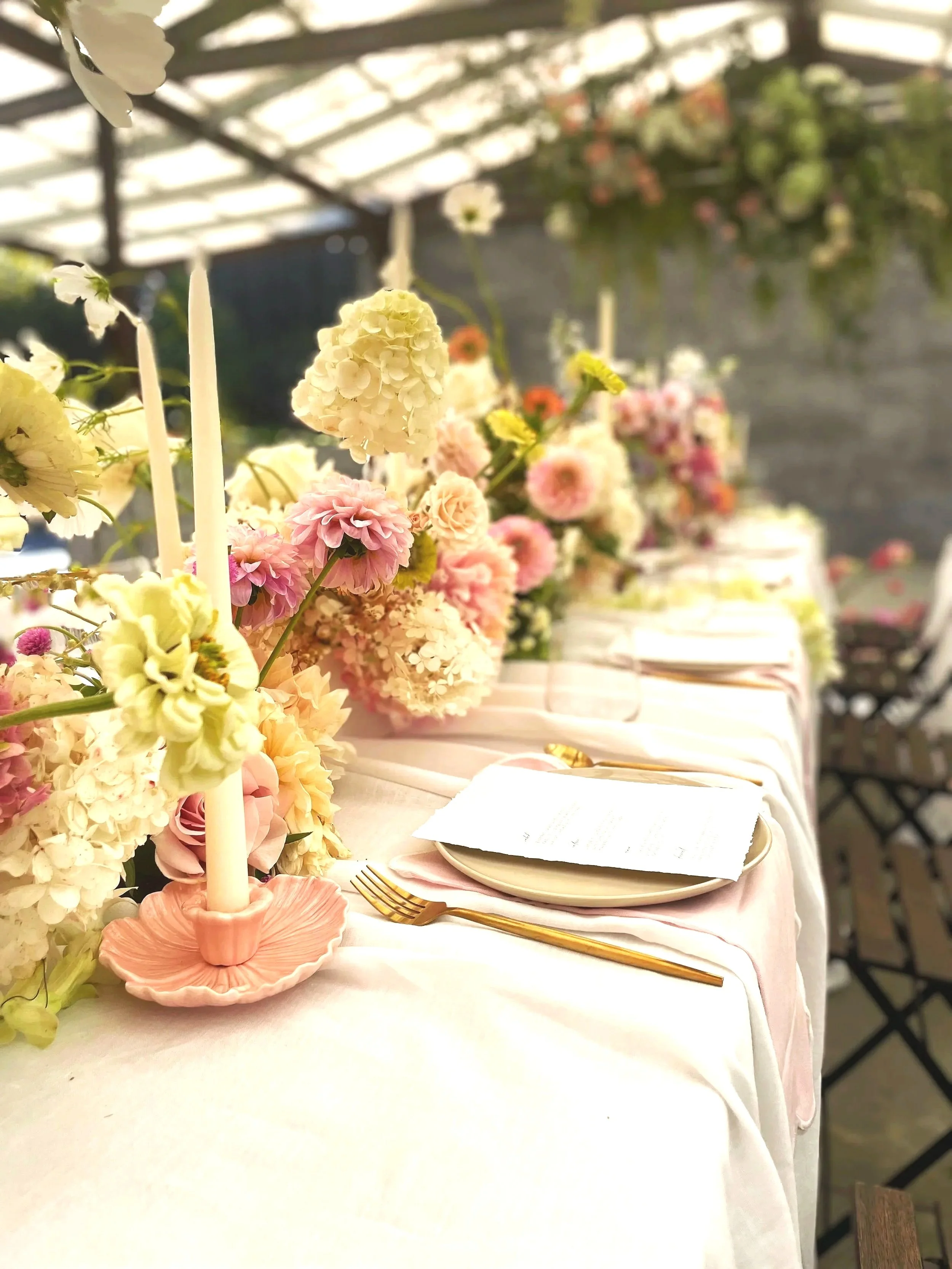 A beautifully decorated table with pink and white flowers, gold utensils, a candle holder, and a menu, set for a formal event or wedding.