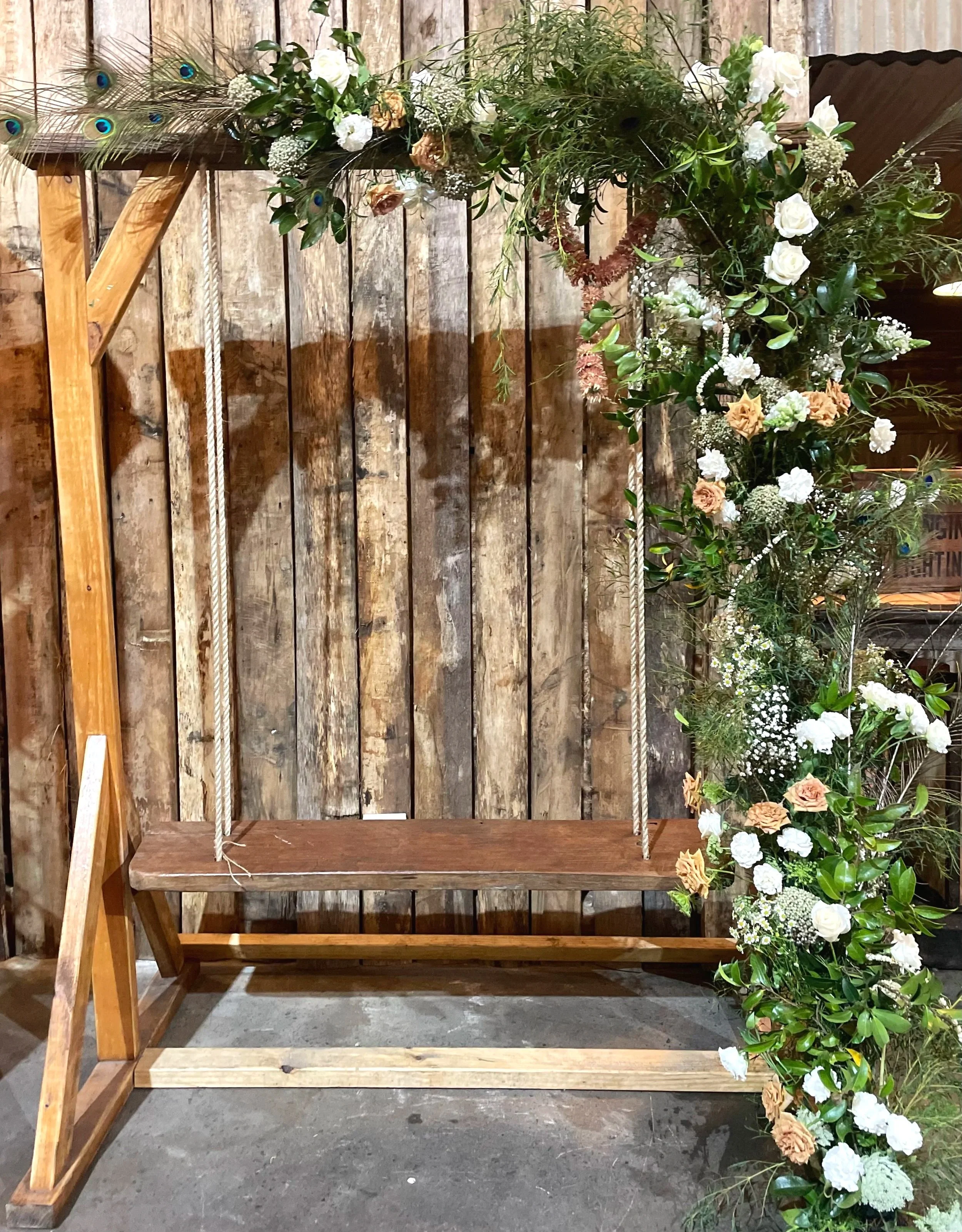 A rustic wooden swing decorated with a wedding garland of white and peach flowers, green leaves, and peacock feathers, set against a weathered wooden wall background.